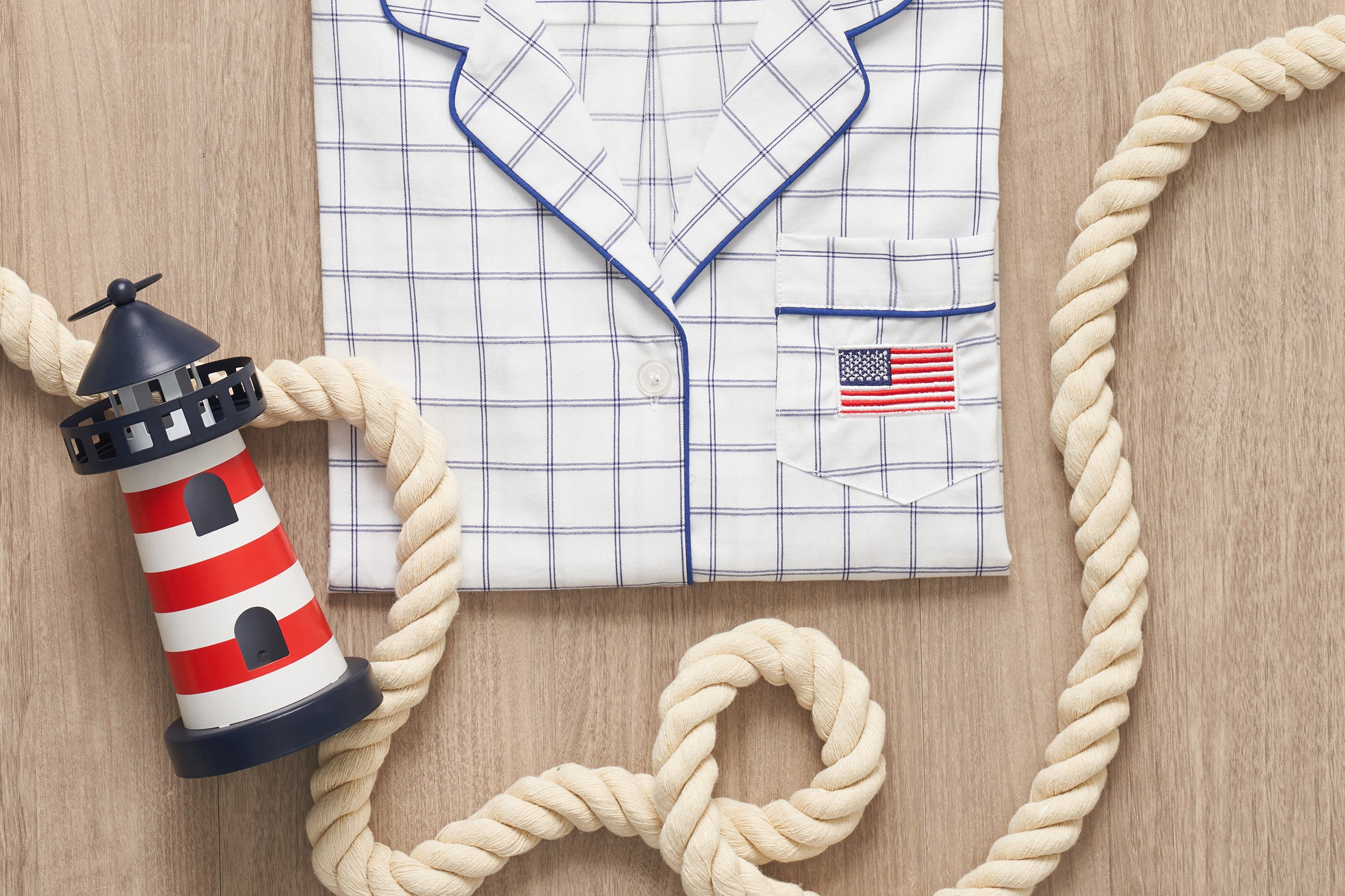 Folded white plaid pajamas with blue trim and an American flag patch on the pocket, next to a toy lighthouse and a thick nautical rope on a wooden surface.