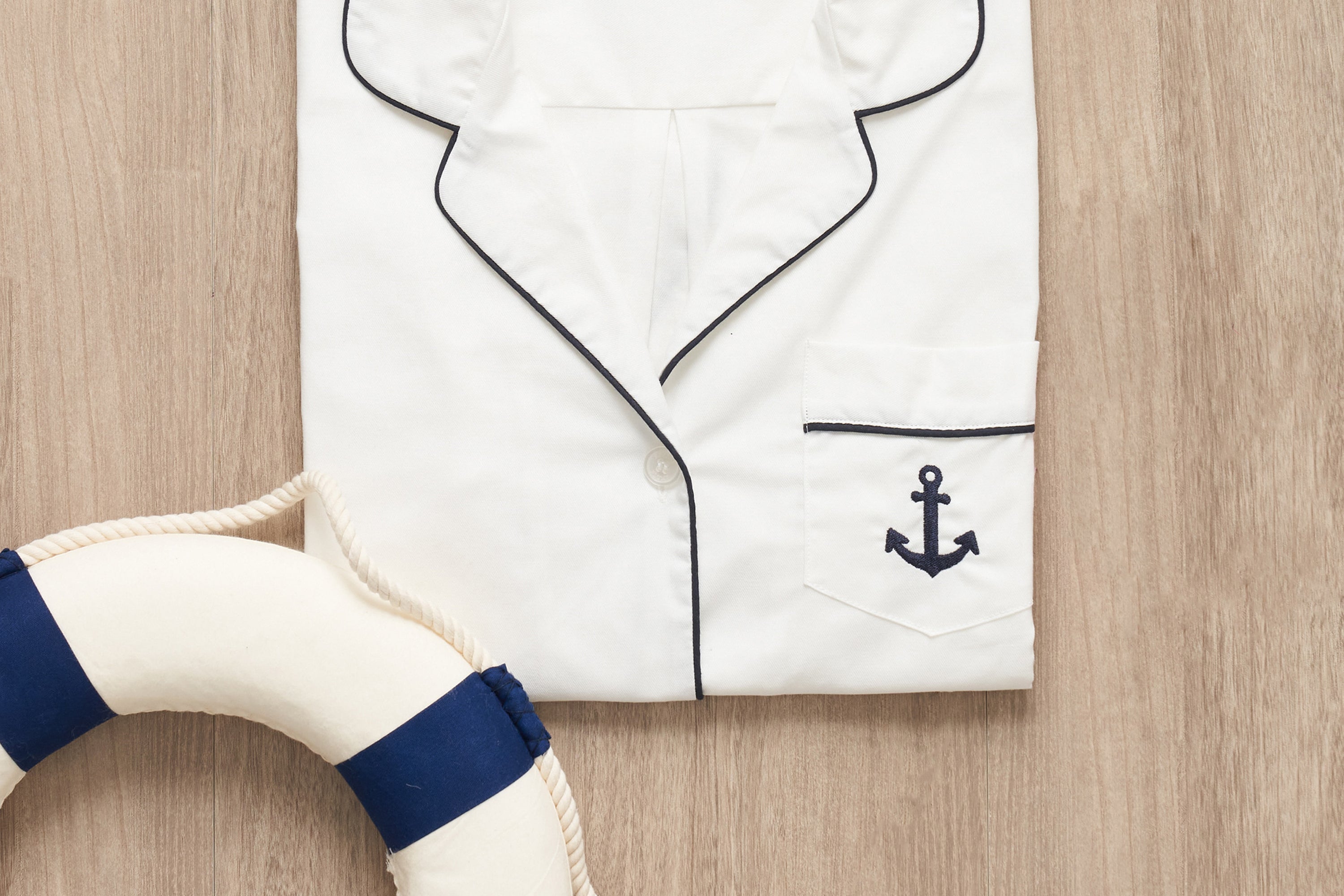 A folded white pajama top with navy piping and an anchor emblem on the pocket, placed on a wooden surface next to a white and blue lifebuoy.