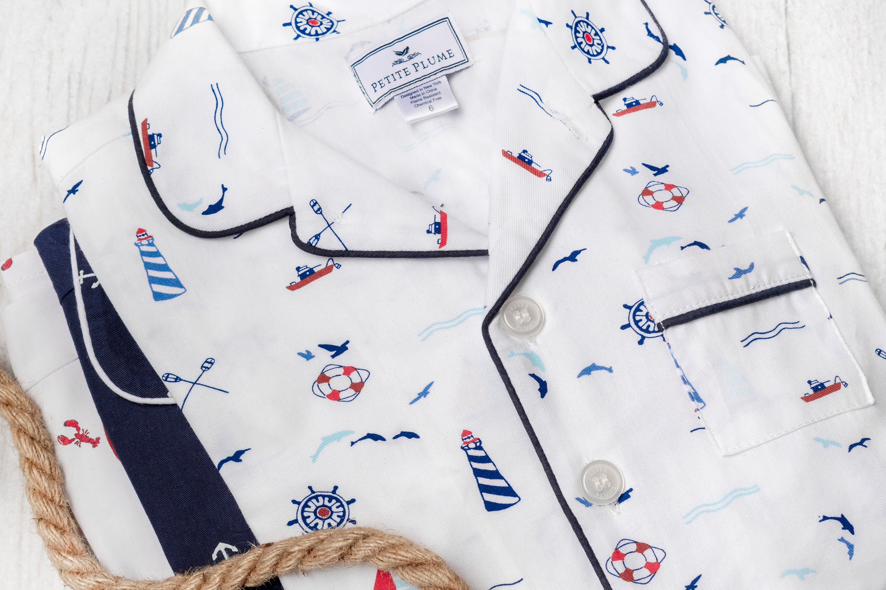 White pajamas with a nautical theme, featuring prints of lighthouses, sailboats, life preservers, anchors, and seagulls, neatly folded with a rope detail nearby.
