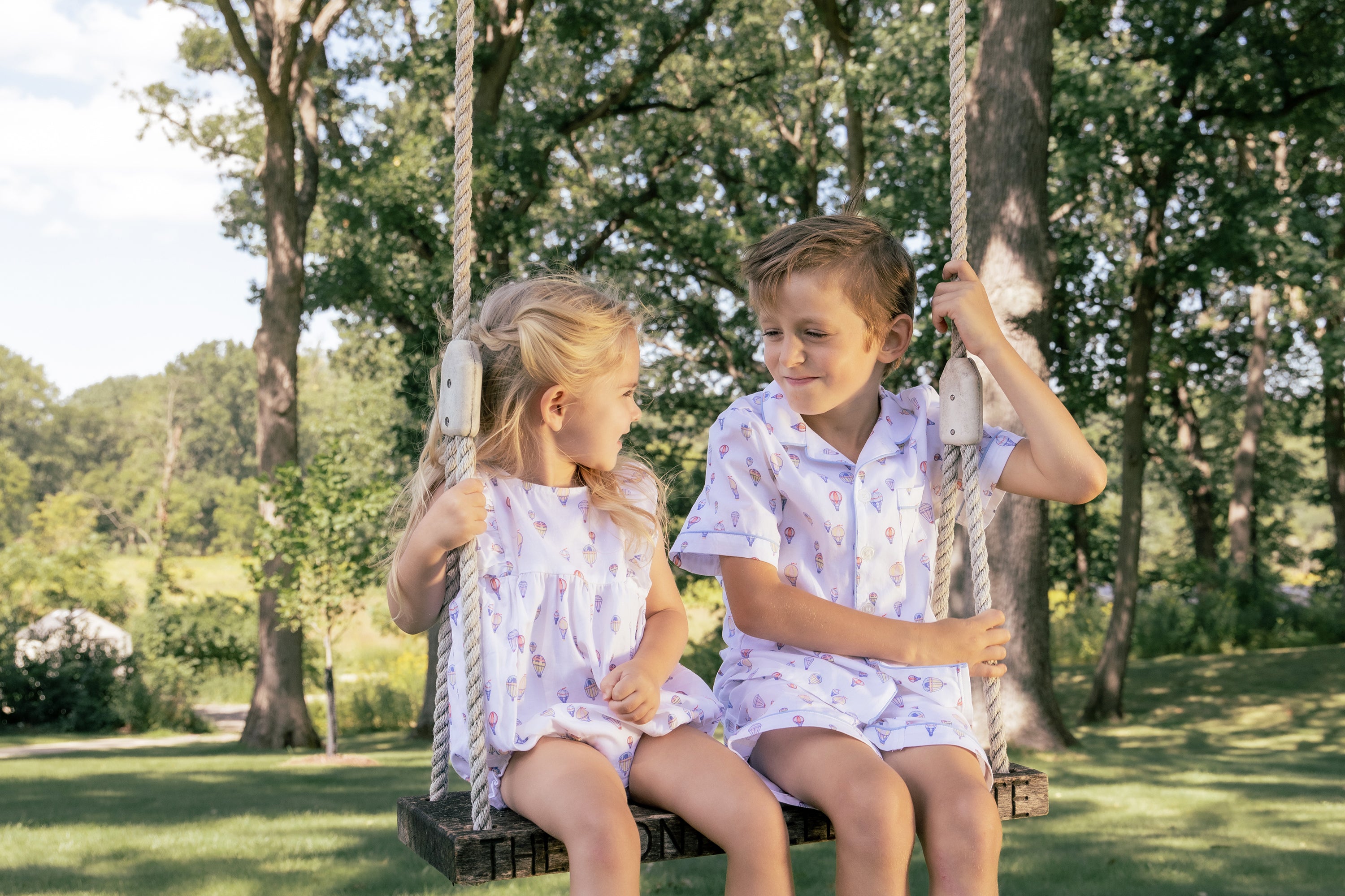 Two young children, a girl and a boy, sit together on a wooden swing outdoors, smiling at each other. They are wearing matching white clothes with colorful patterns. Trees and greenery surround them.