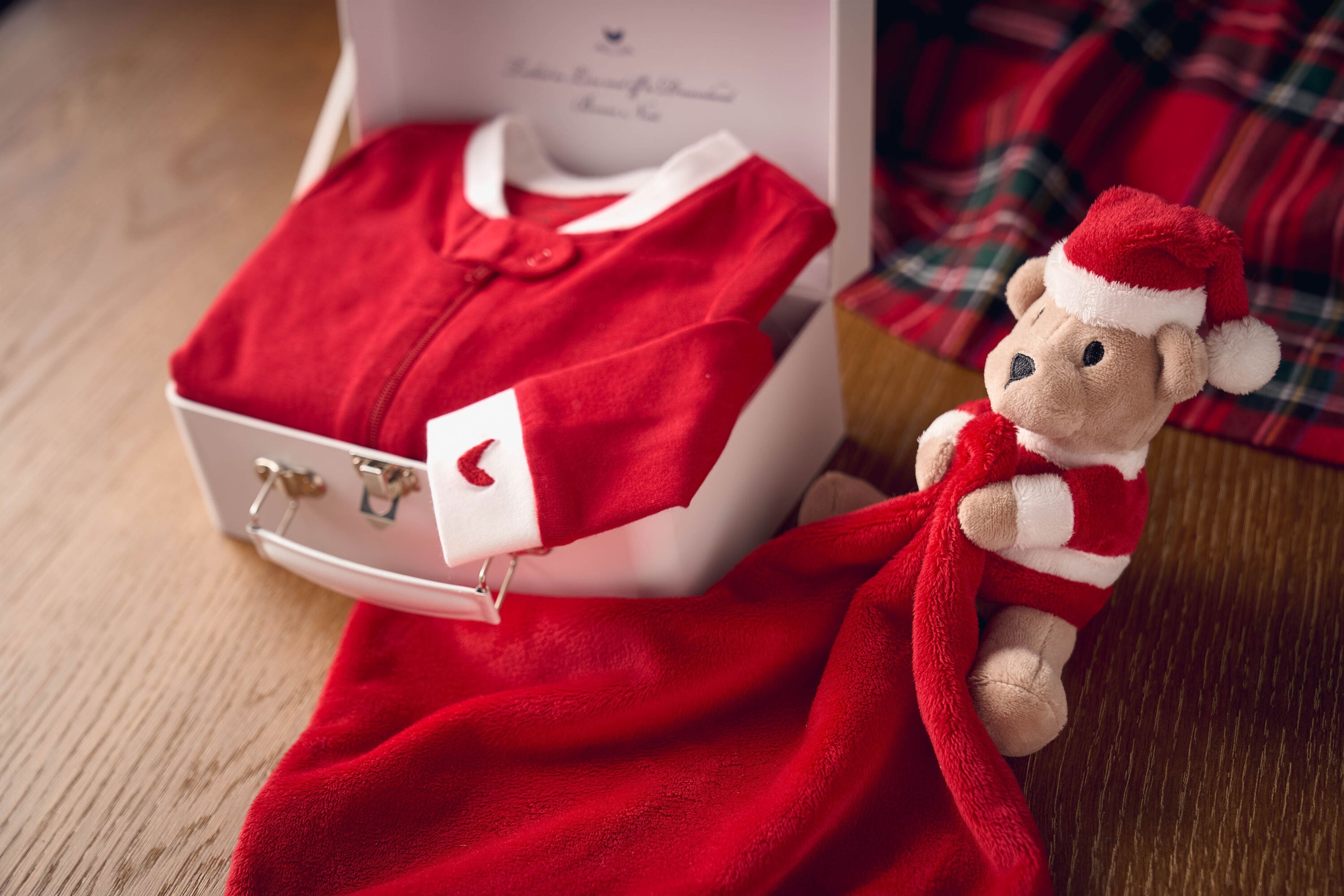 A red baby outfit in a white box lies on a wooden surface next to a stuffed teddy bear dressed in a red and white Santa outfit, holding a red blanket. A plaid fabric is in the background.