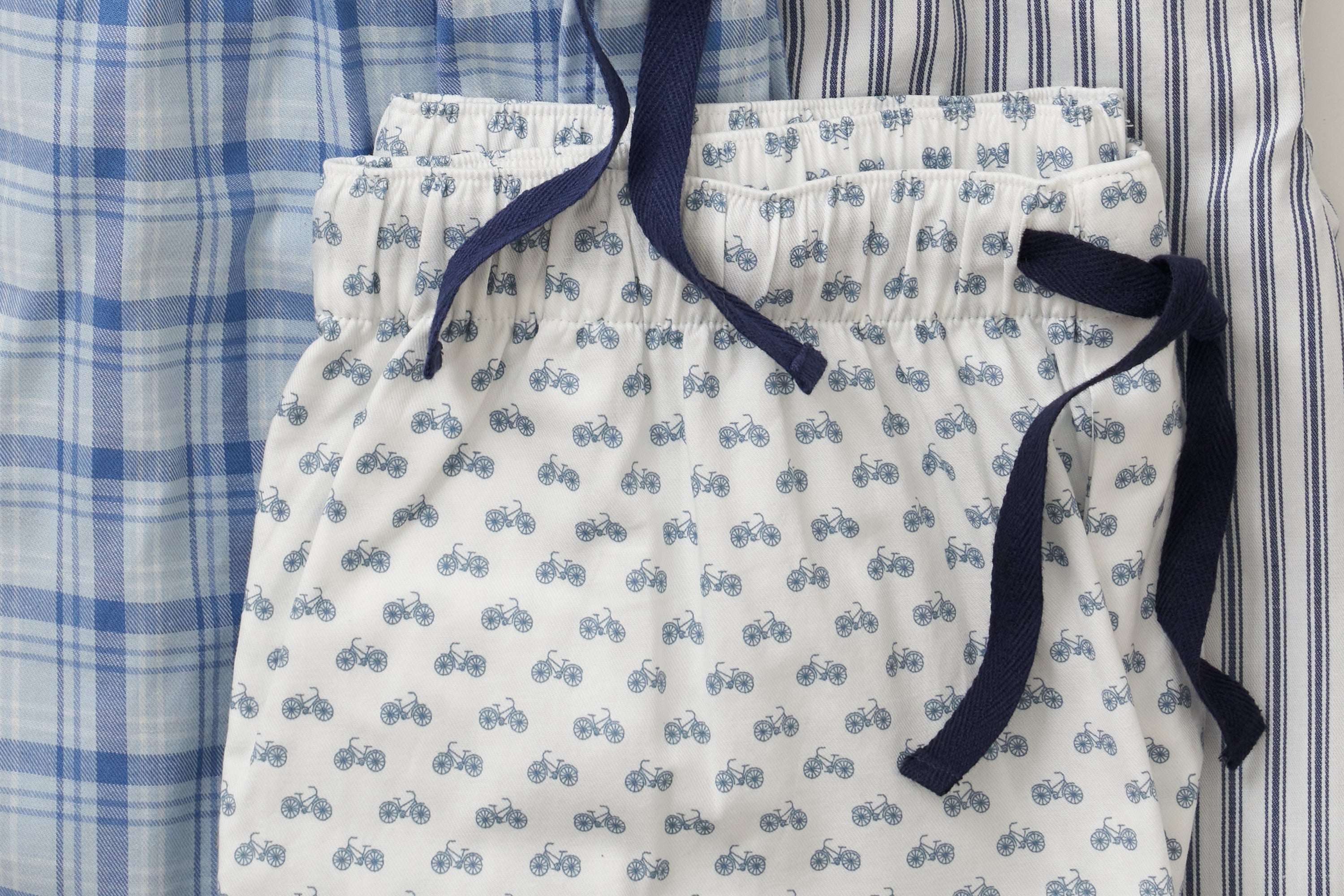 Three pairs of pajama pants with different patterns: blue plaid, blue stripes, and white with blue bicycles, all with drawstrings.