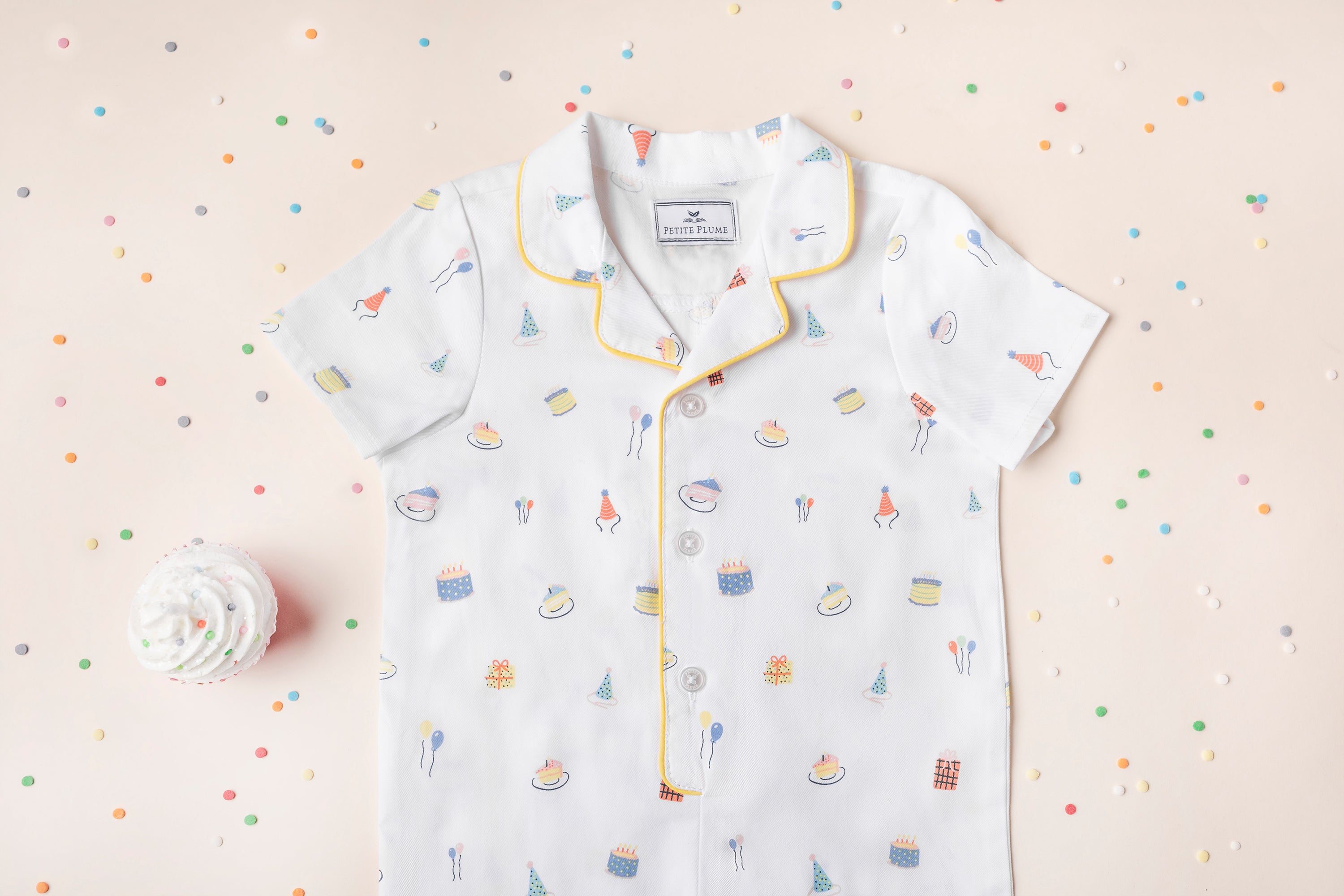 A white short-sleeve shirt with colorful party-themed prints, yellow piping on the collar and buttons, is laid flat on a pastel background with scattered confetti and a cupcake.