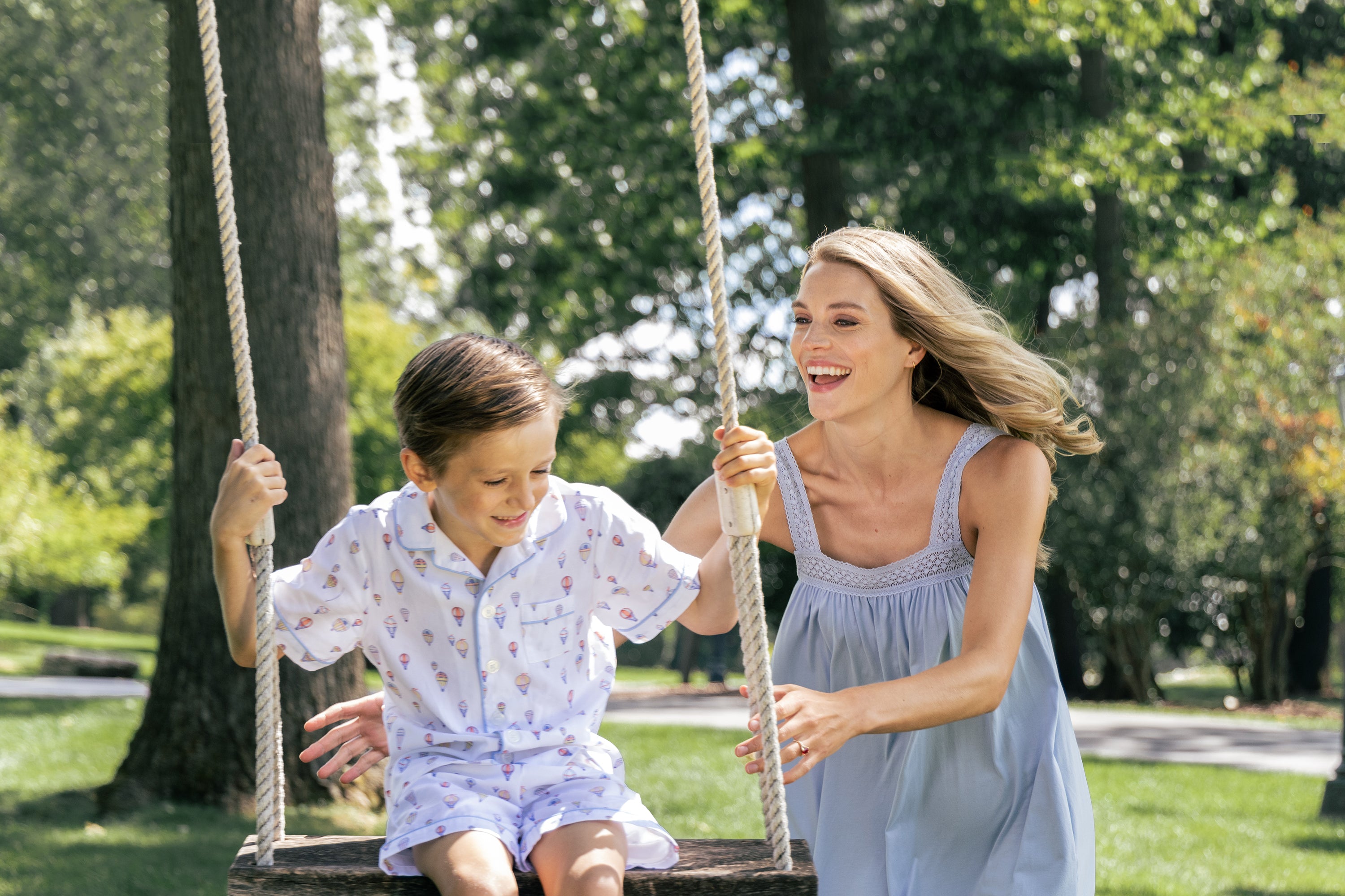 A woman pushes a smiling young boy on a wooden swing in a sunny park, surrounded by green grass and trees. Both appear happy and are enjoying the outdoors.