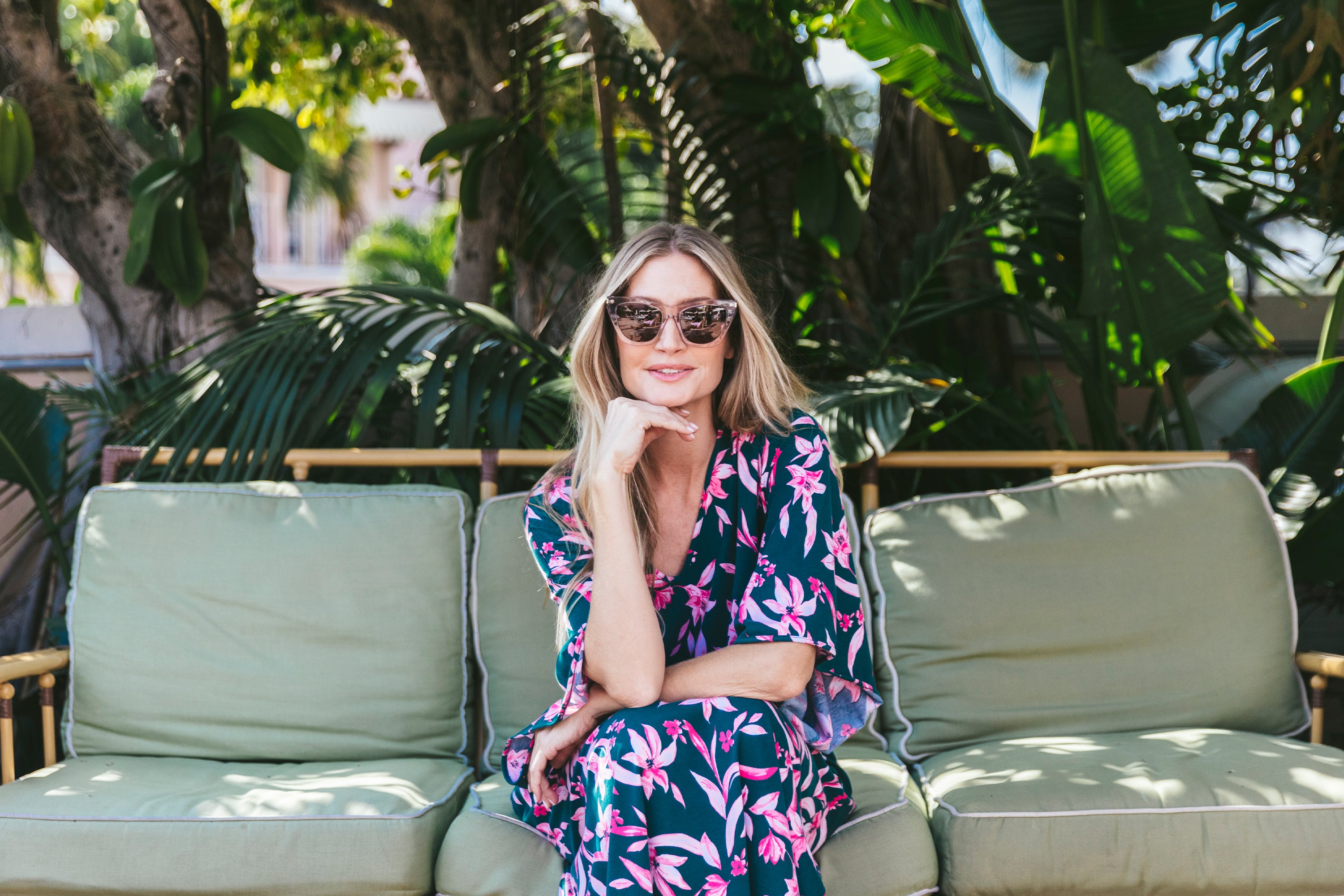 A woman in sunglasses and a floral dress sits on a green outdoor couch, surrounded by lush greenery. She rests her chin on her hand and smiles at the camera on a sunny day.