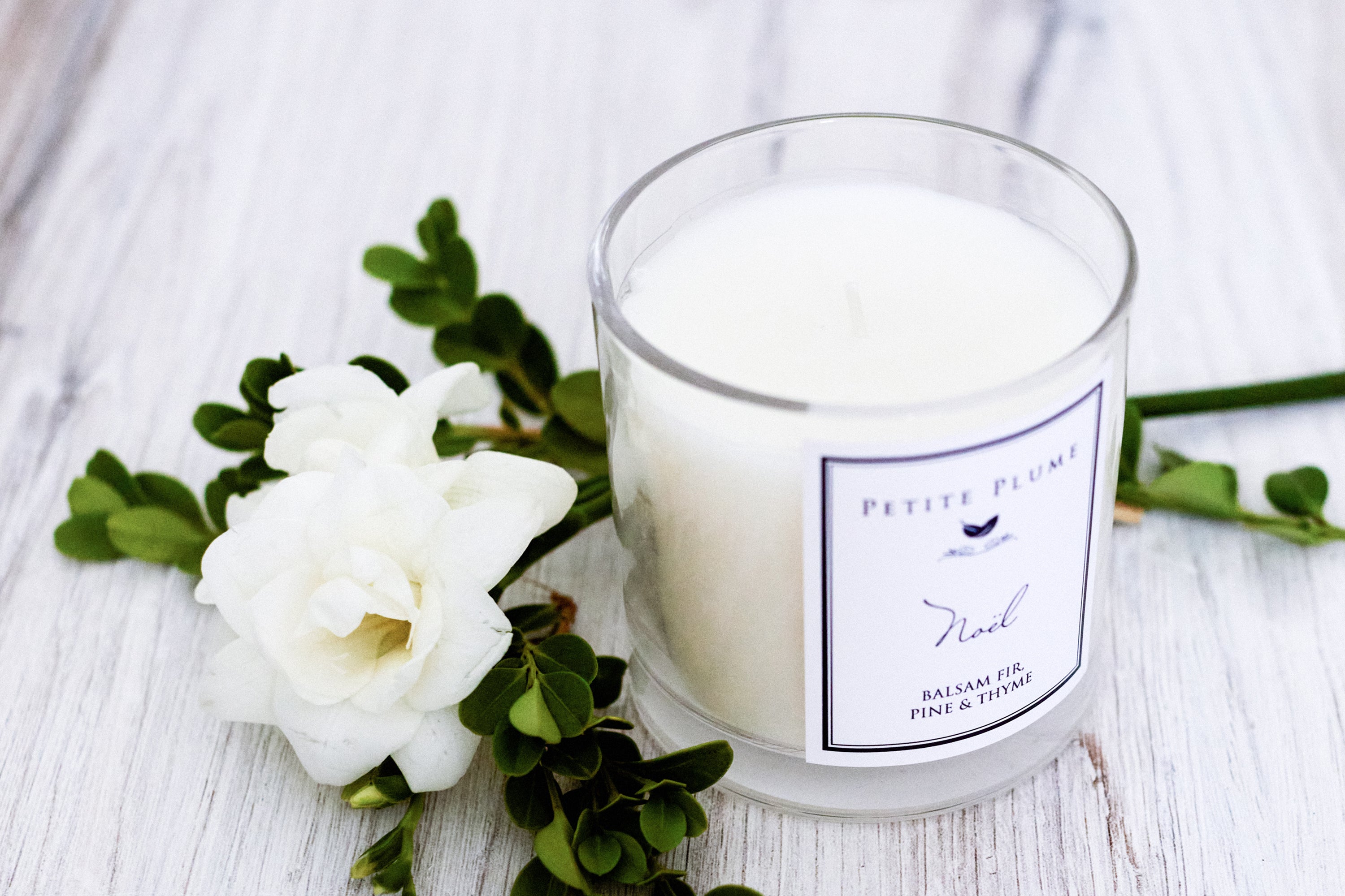 A white candle in a glass jar labeled Petite Plume sits on a light wooden surface next to white flowers and green leaves.