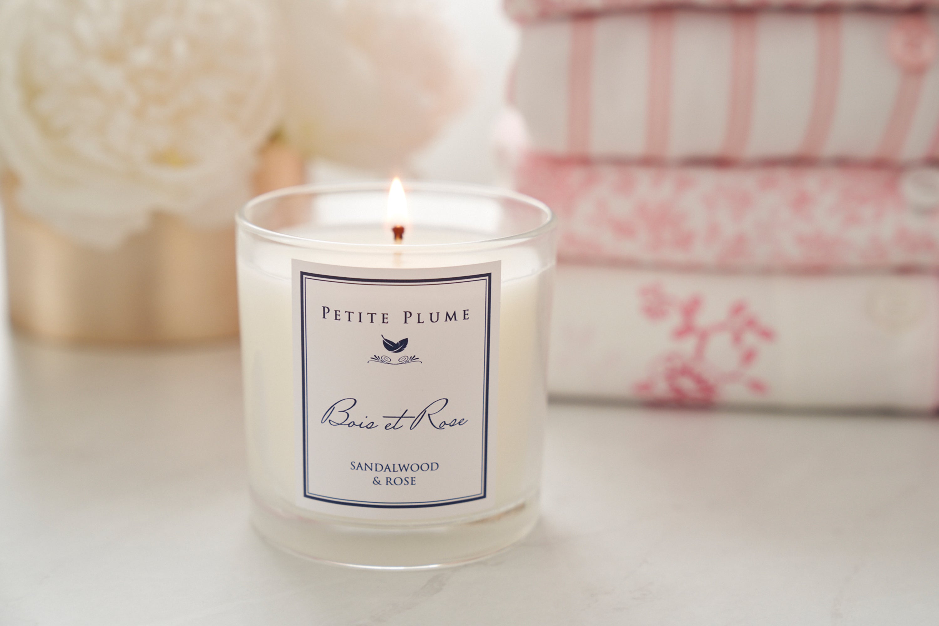 A lit candle labeled Petite Plume, Bois et Rose, Sandalwood & Rose sits on a white surface, with soft pink and white folded fabrics and a white flower in the background.