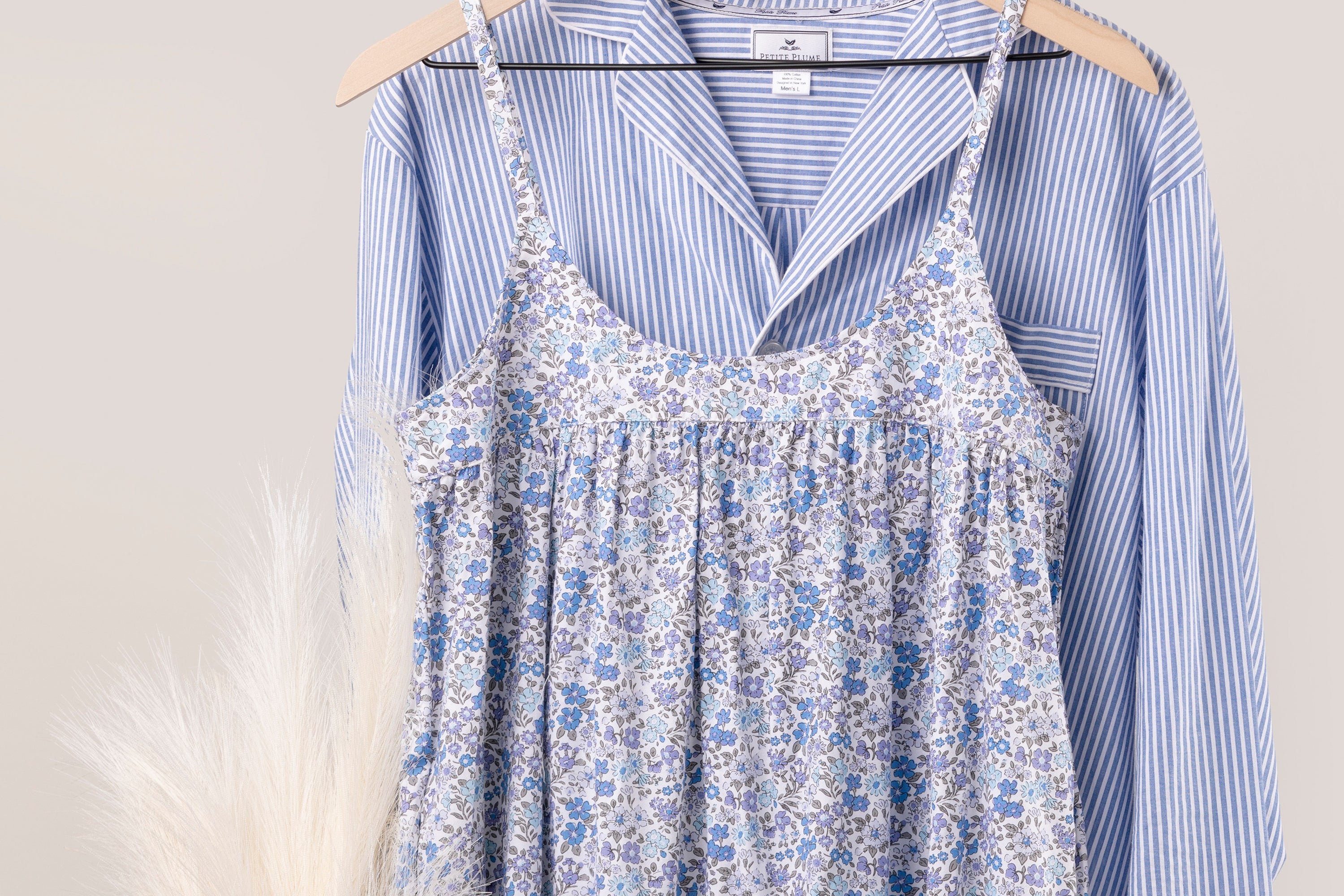 A blue floral dress on a wooden hanger is layered over a blue and white striped button-up shirt, also on a hanger. Pampas grass is partially visible on the left side.