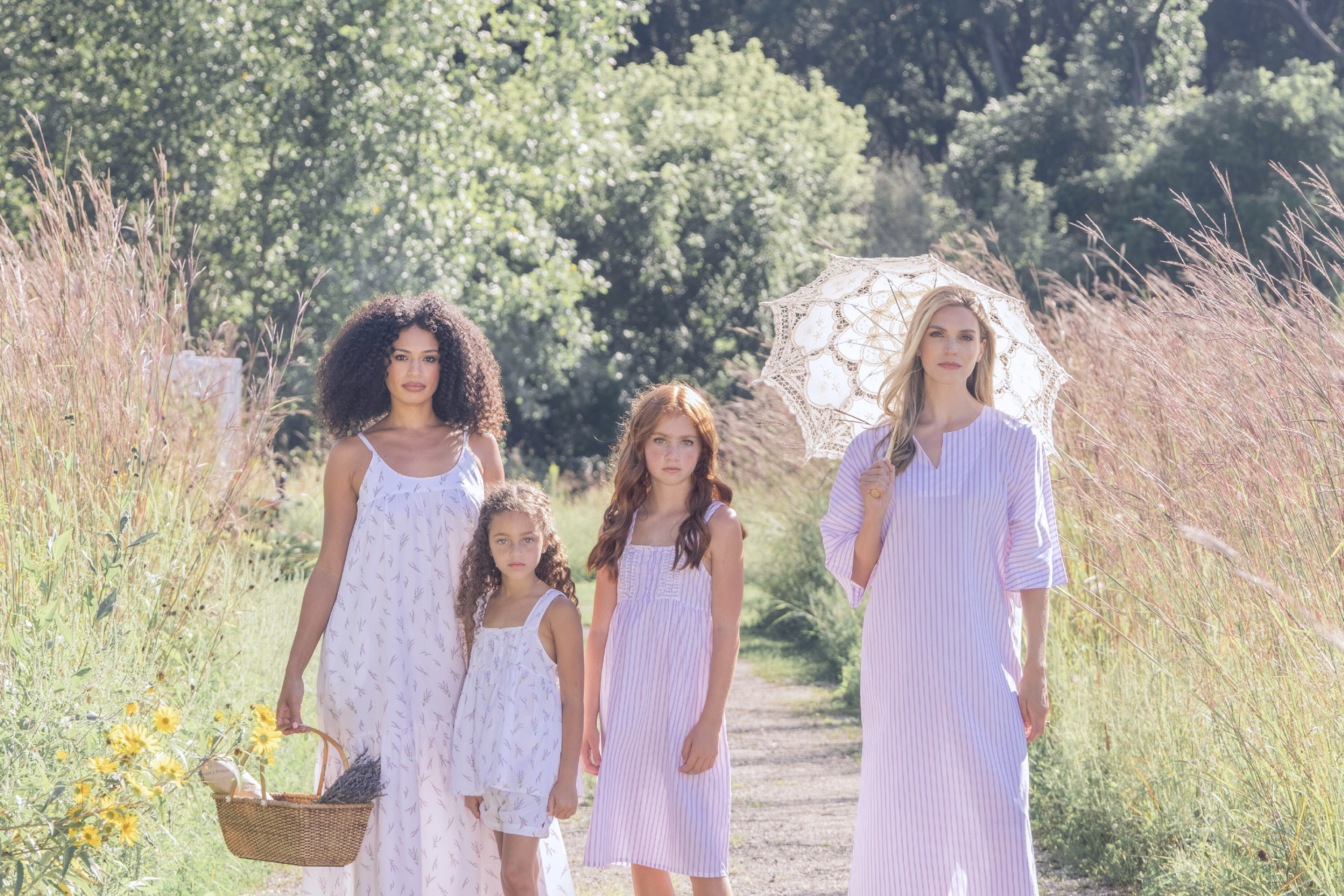 Four women and girls in light, summery dresses stand on a sunlit path in tall grass. One holds a basket, another holds a lace parasol. The scene is bright, calm, and natural.