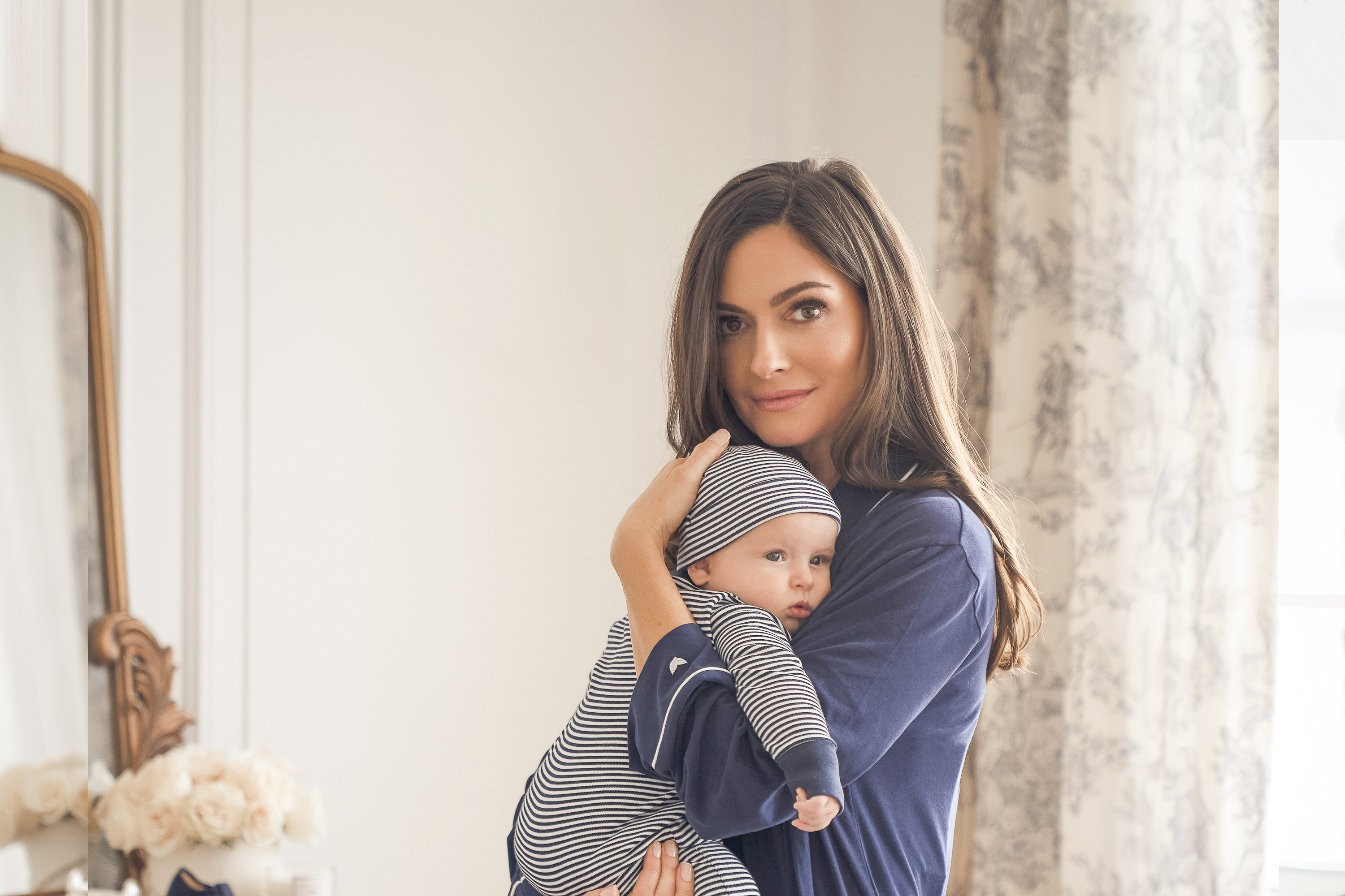 A woman in a blue top gently holds a baby dressed in a striped outfit and hat. They are standing indoors near a window with floral curtains and a vase of flowers on a dresser in the background.