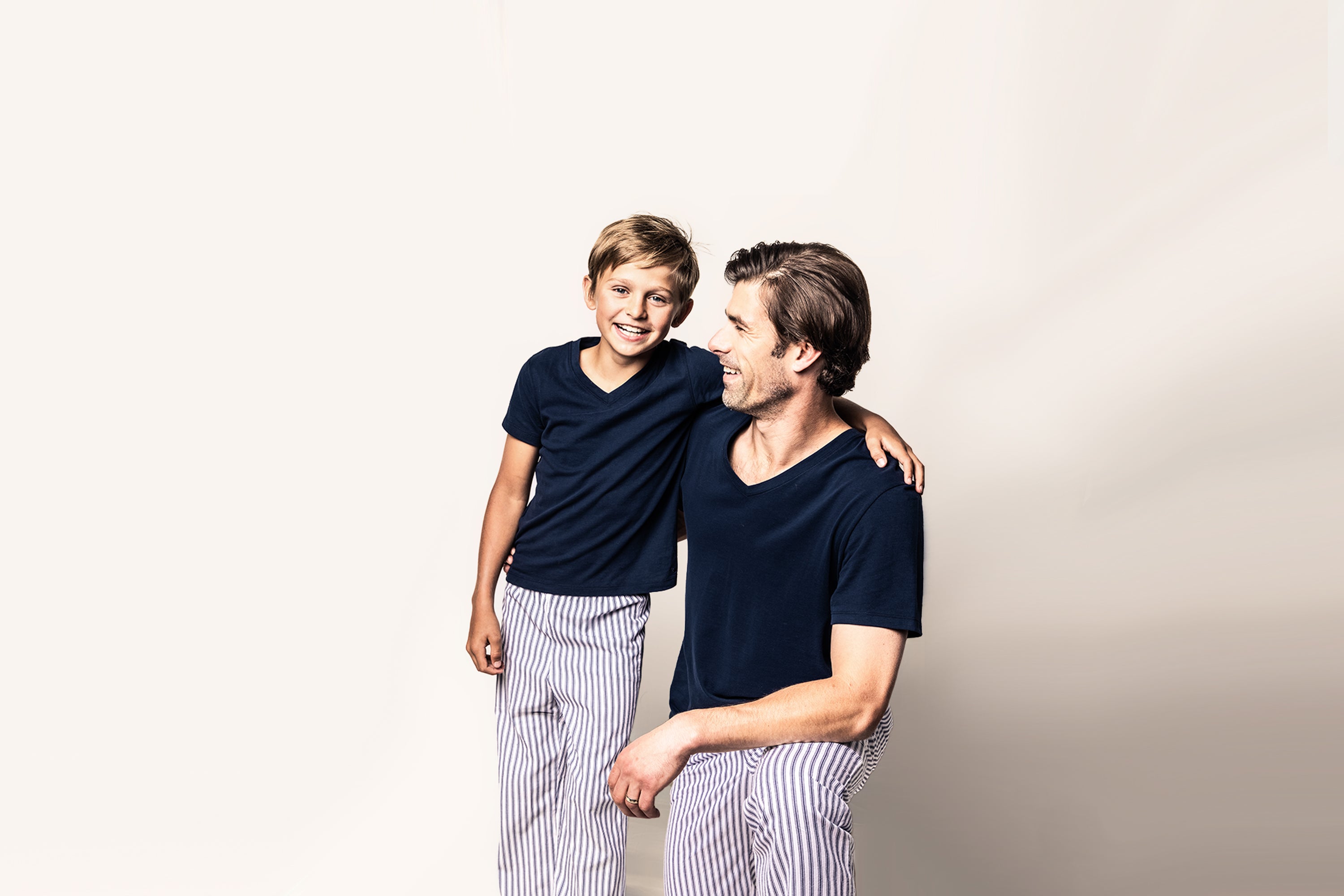 A smiling boy and a man, both wearing matching navy t-shirts and striped pants, stand together against a light background. The boy has his arm around the mans shoulder.