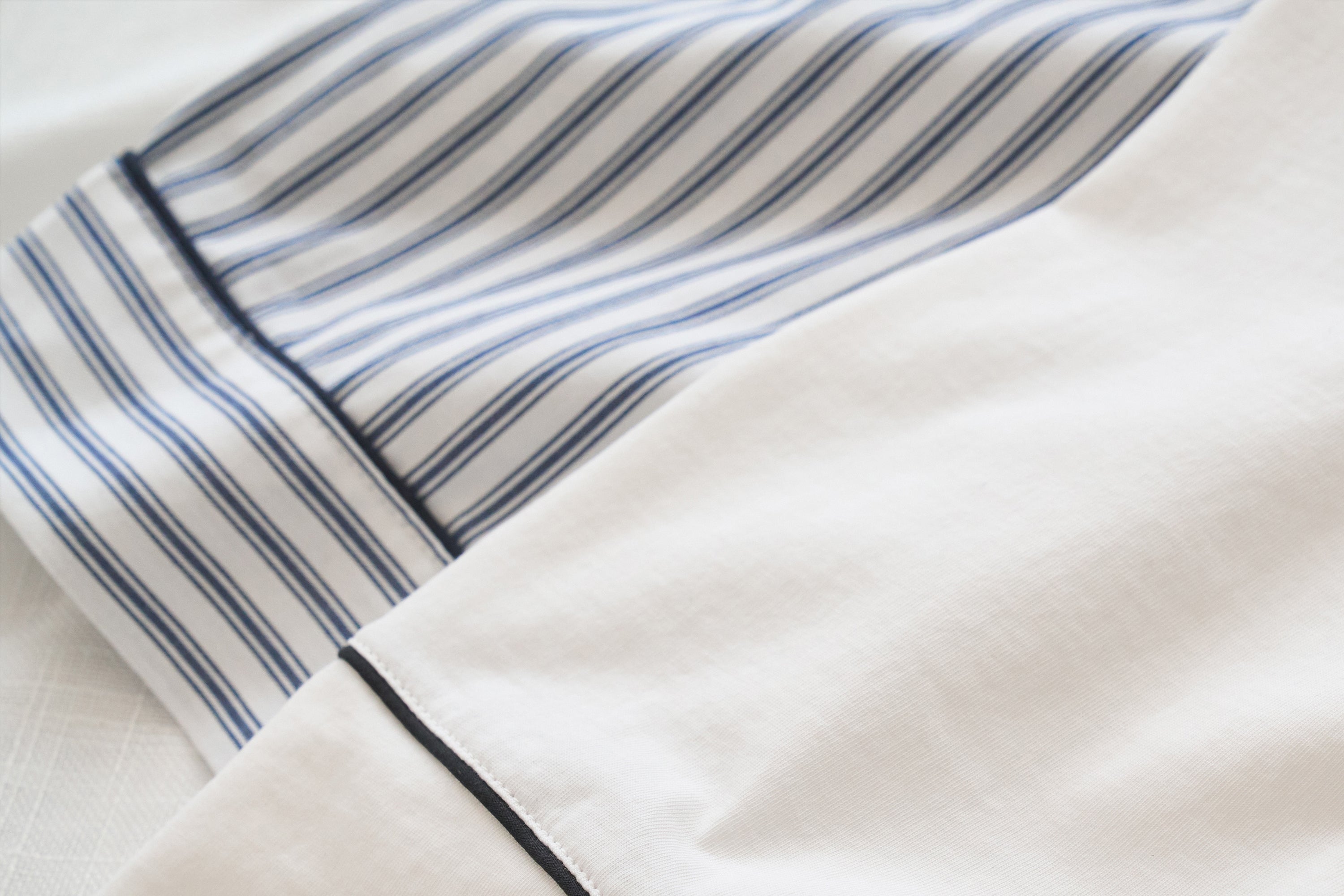 Close-up of a white fabric with a blue and white striped cuff, accented by a thin black piping detail. The material appears smooth and neatly pressed, suggesting it is part of a dress shirt or bedding.