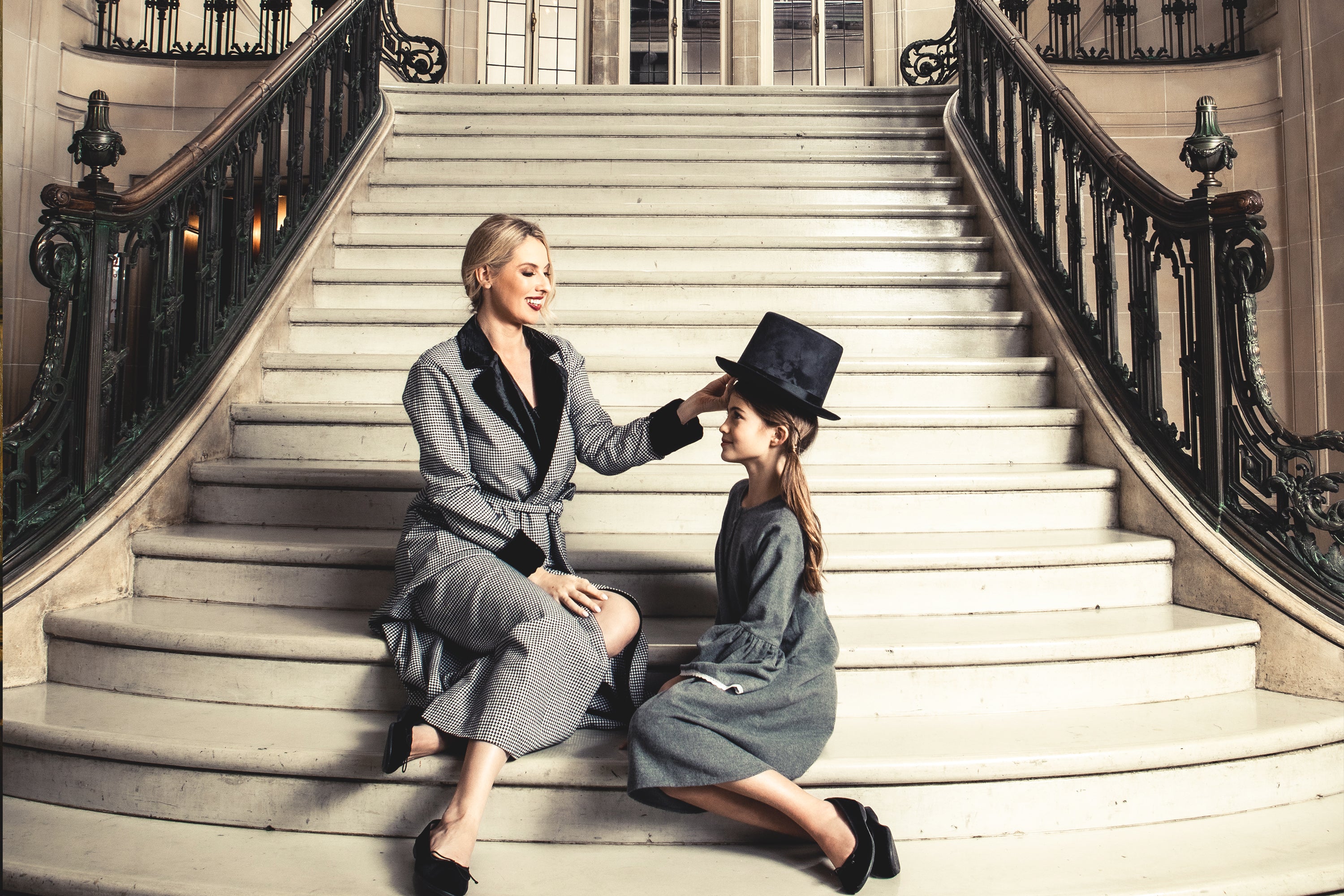 A woman sitting on grand stairs smiles as she places a top hat on a young girl kneeling beside her. Both are wearing grey coats in an elegant, ornate interior.