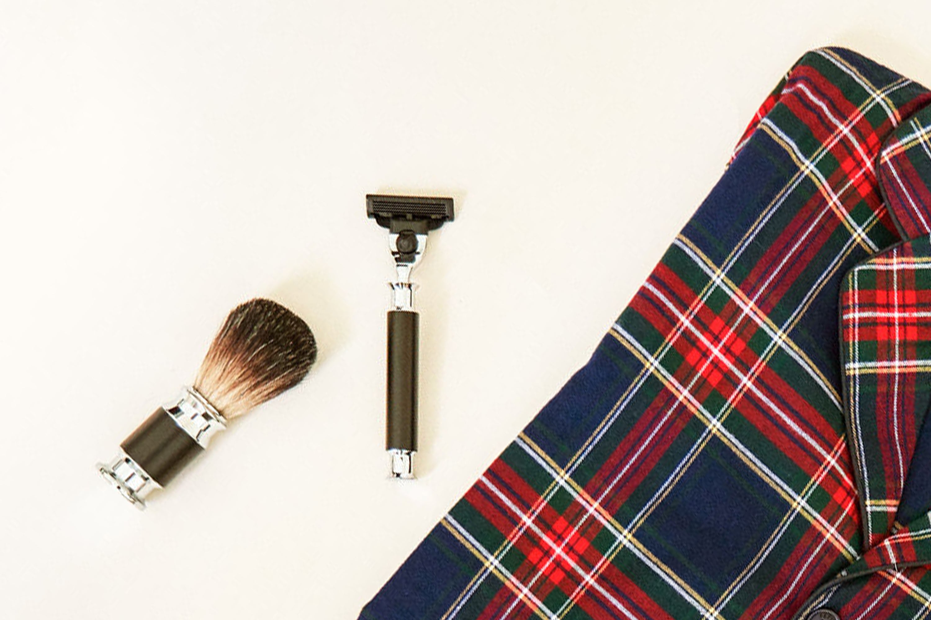 A shaving brush, safety razor, and a folded red and blue plaid shirt are arranged neatly on a white background.