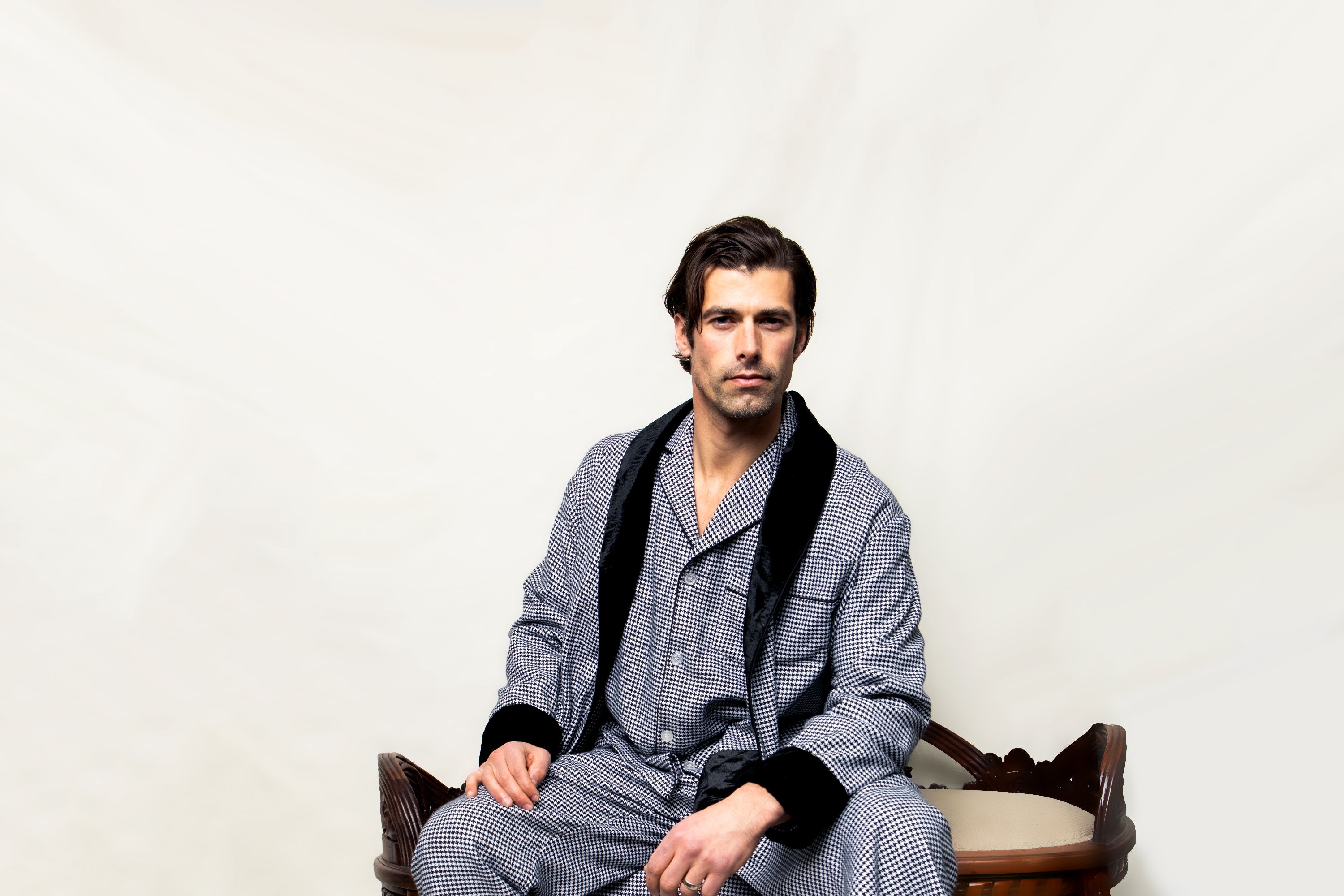 A man with dark hair wearing a patterned gray robe sits on a wooden chair with a neutral background, looking directly at the camera with a serious expression.