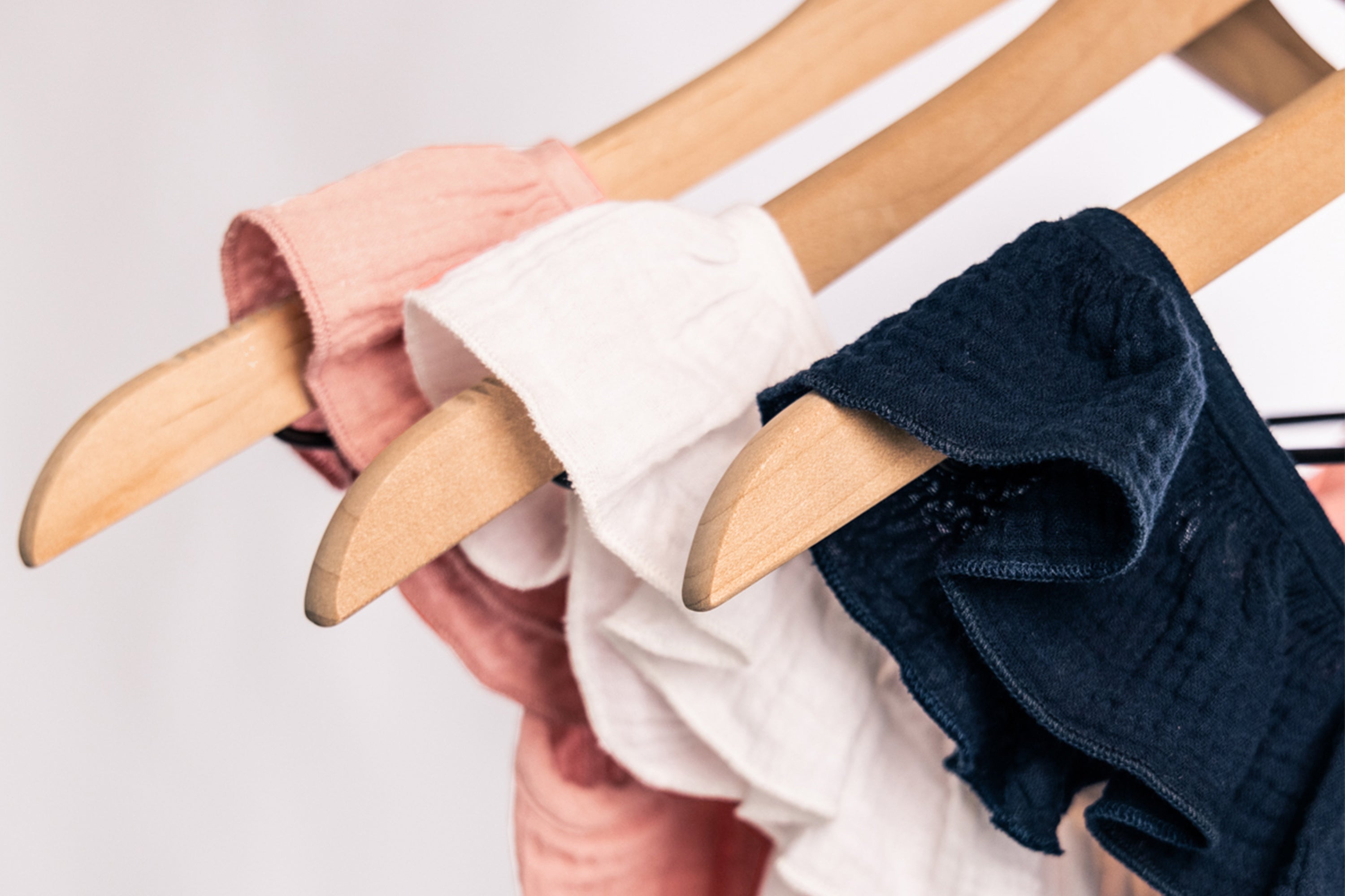 Three pairs of cotton underwear in pink, white, and navy hang neatly on wooden hangers against a soft, light background.
