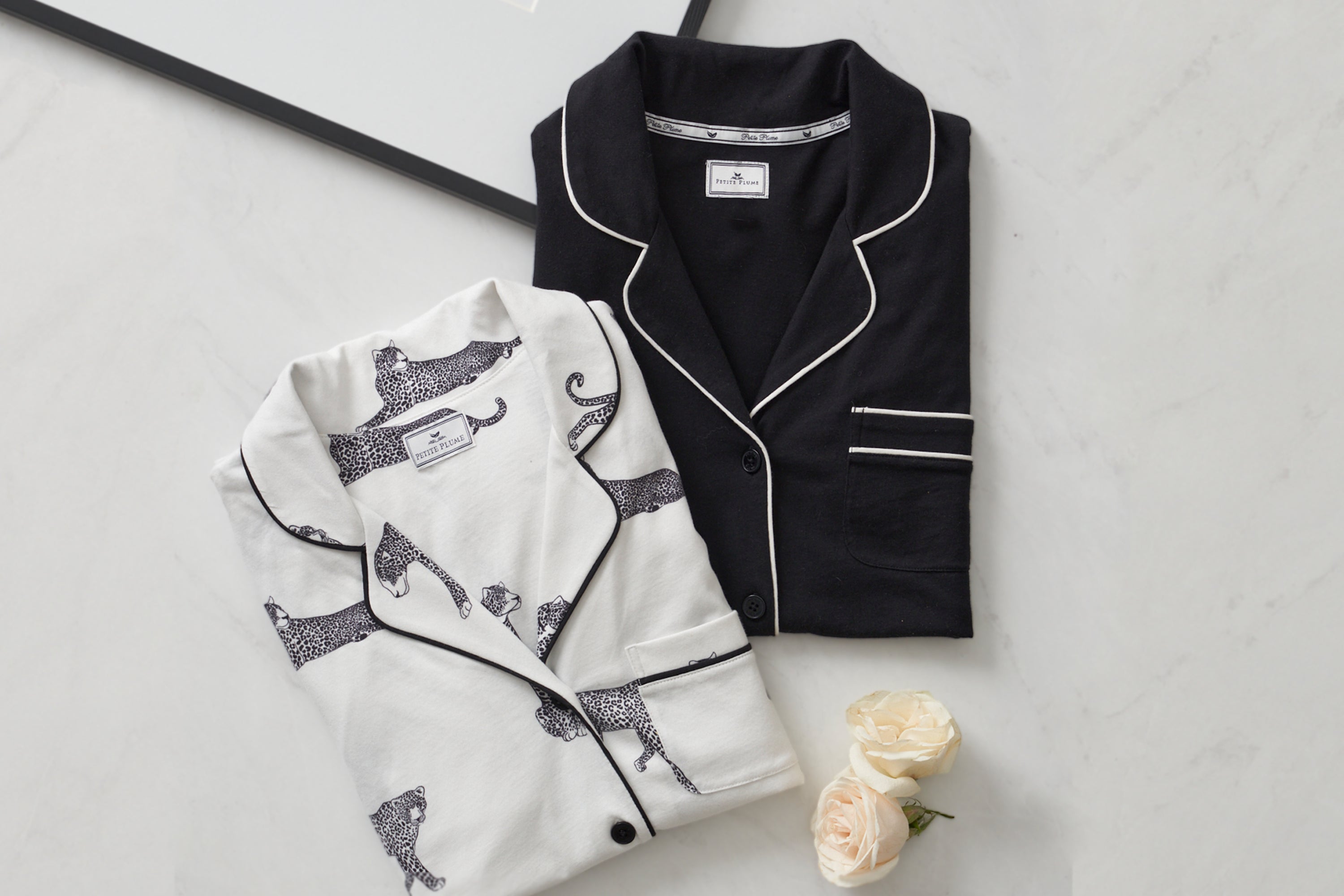 Two folded pajama tops on a light surface; one is white with a black cheetah print, the other is solid black with white piping. Two ivory roses are placed nearby.