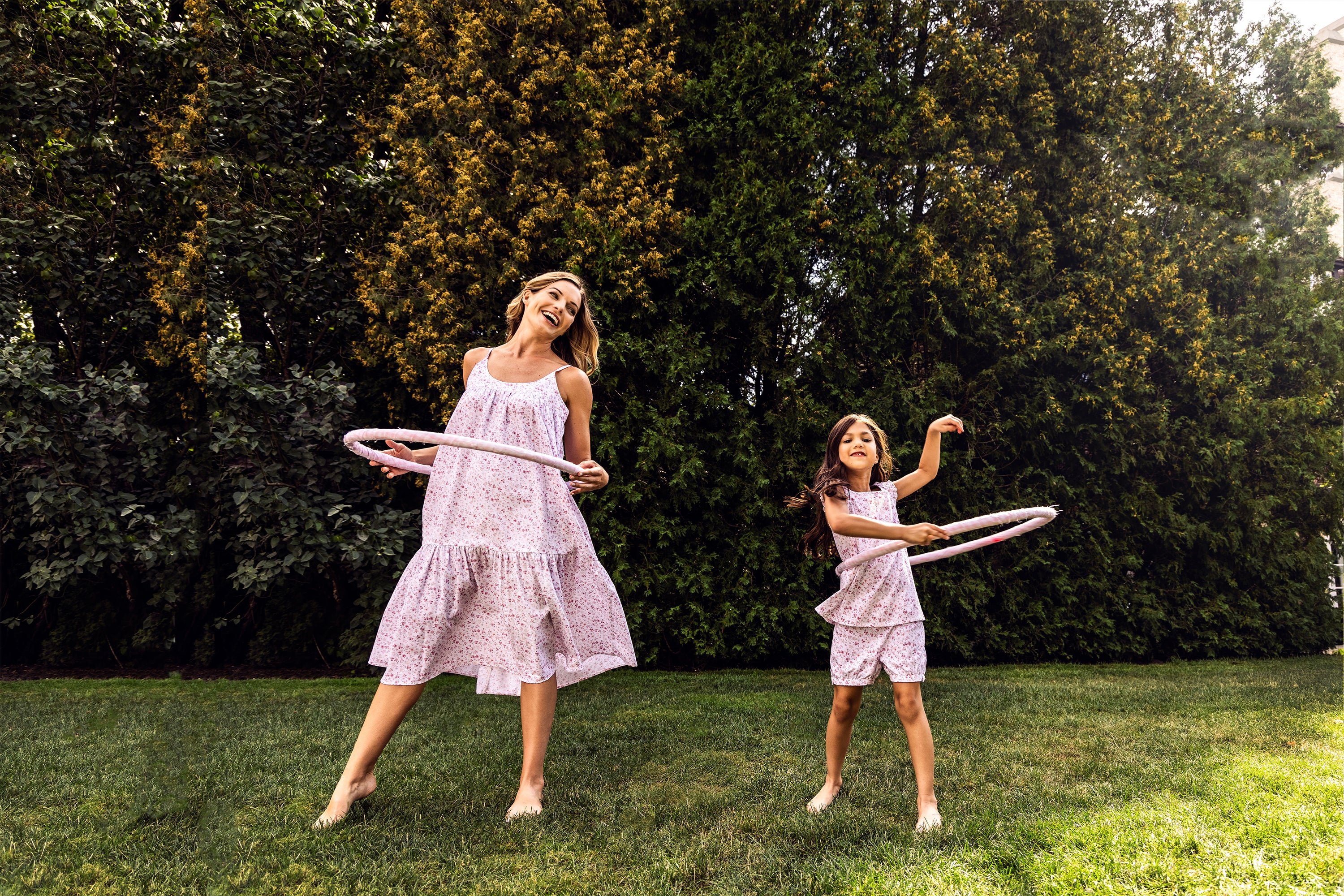 A woman and a young girl wearing matching light pink outfits play with hula hoops on a grassy lawn, smiling and having fun, with tall green shrubs in the background.