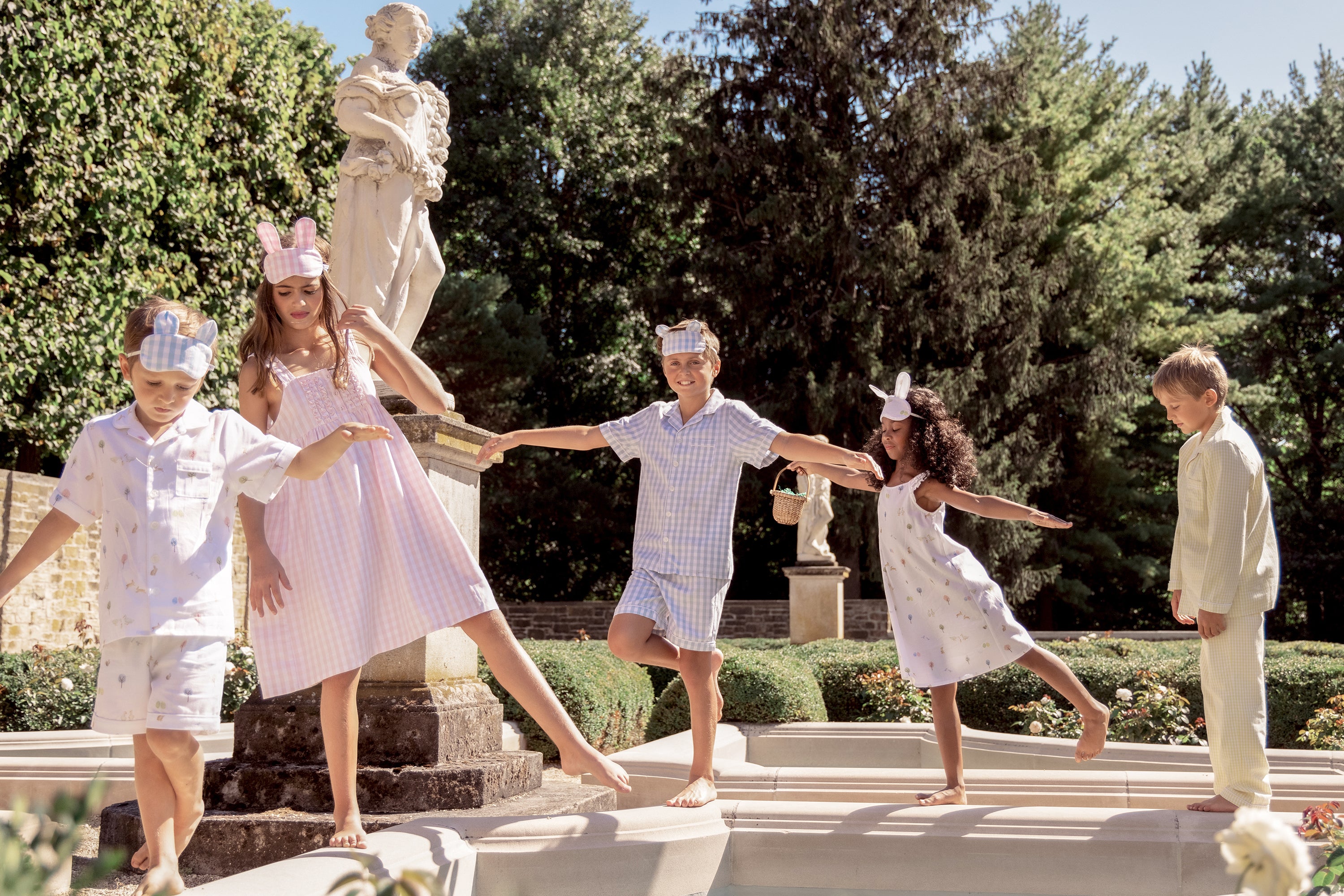 Five children in pastel pajamas and animal ear headbands balance on the edge of a garden fountain. Statues and greenery surround them on a sunny day. They appear playful and joyful.