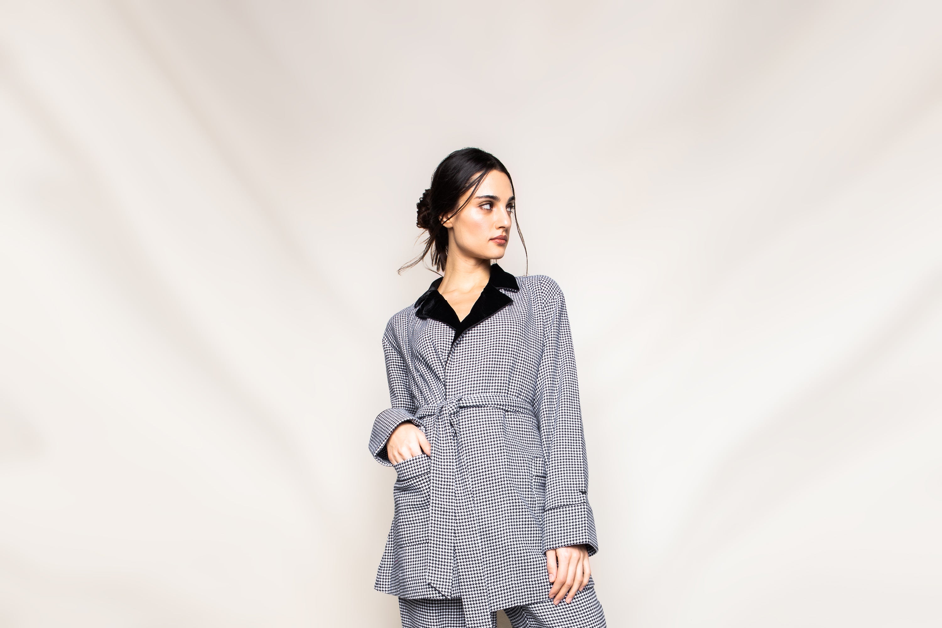 A woman with dark hair styled in a low bun wears a gray checkered suit, looking to the side against a plain, light beige background.