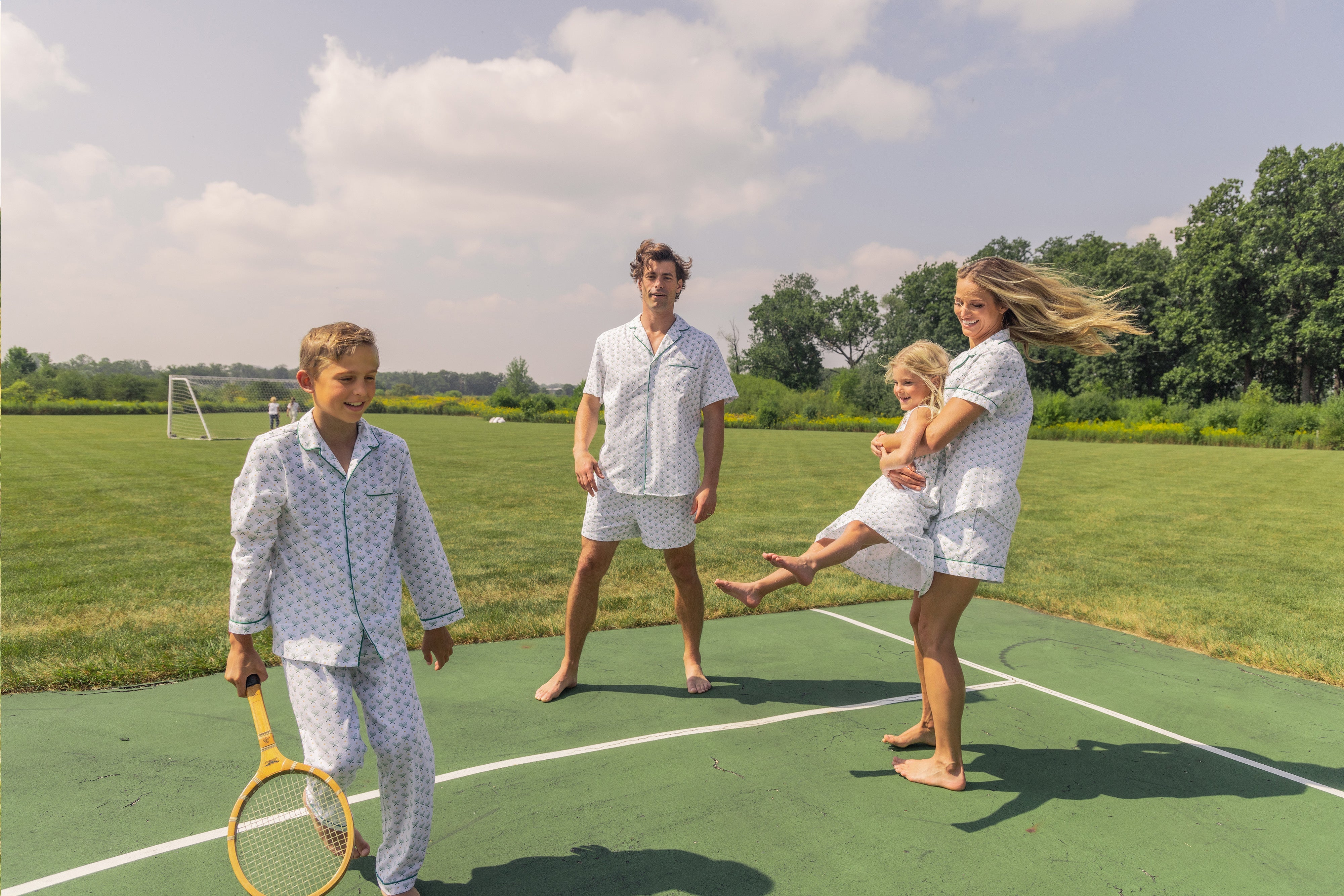 A family in matching pajamas is joyfully playing on a tennis court. The mother holds a young child, the father stands nearby, and a boy walks with a tennis racket. The sky is partly cloudy in the background.