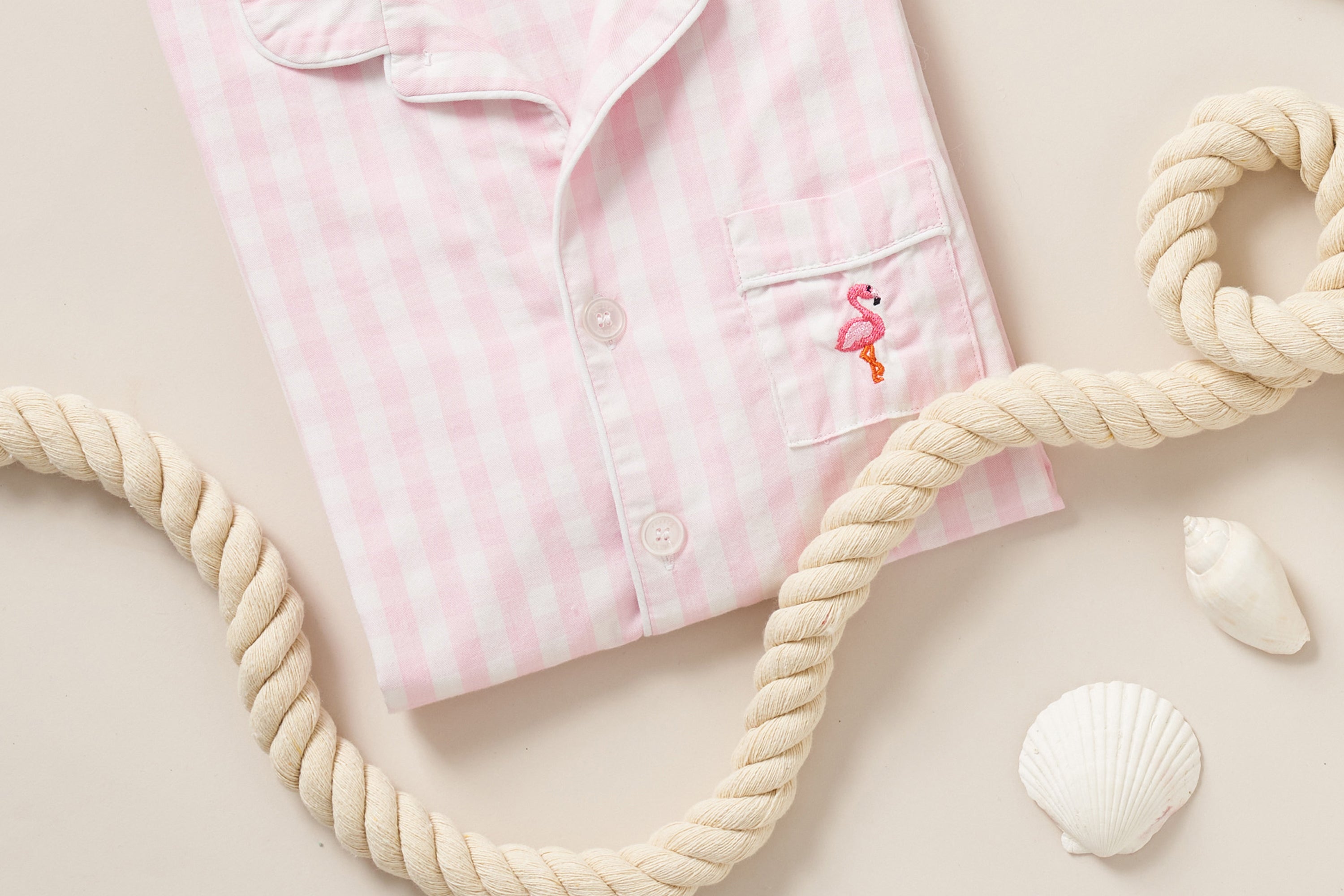 A folded pink and white checkered pajama top with an embroidered flamingo on the pocket, surrounded by a thick rope and white seashells on a beige background.