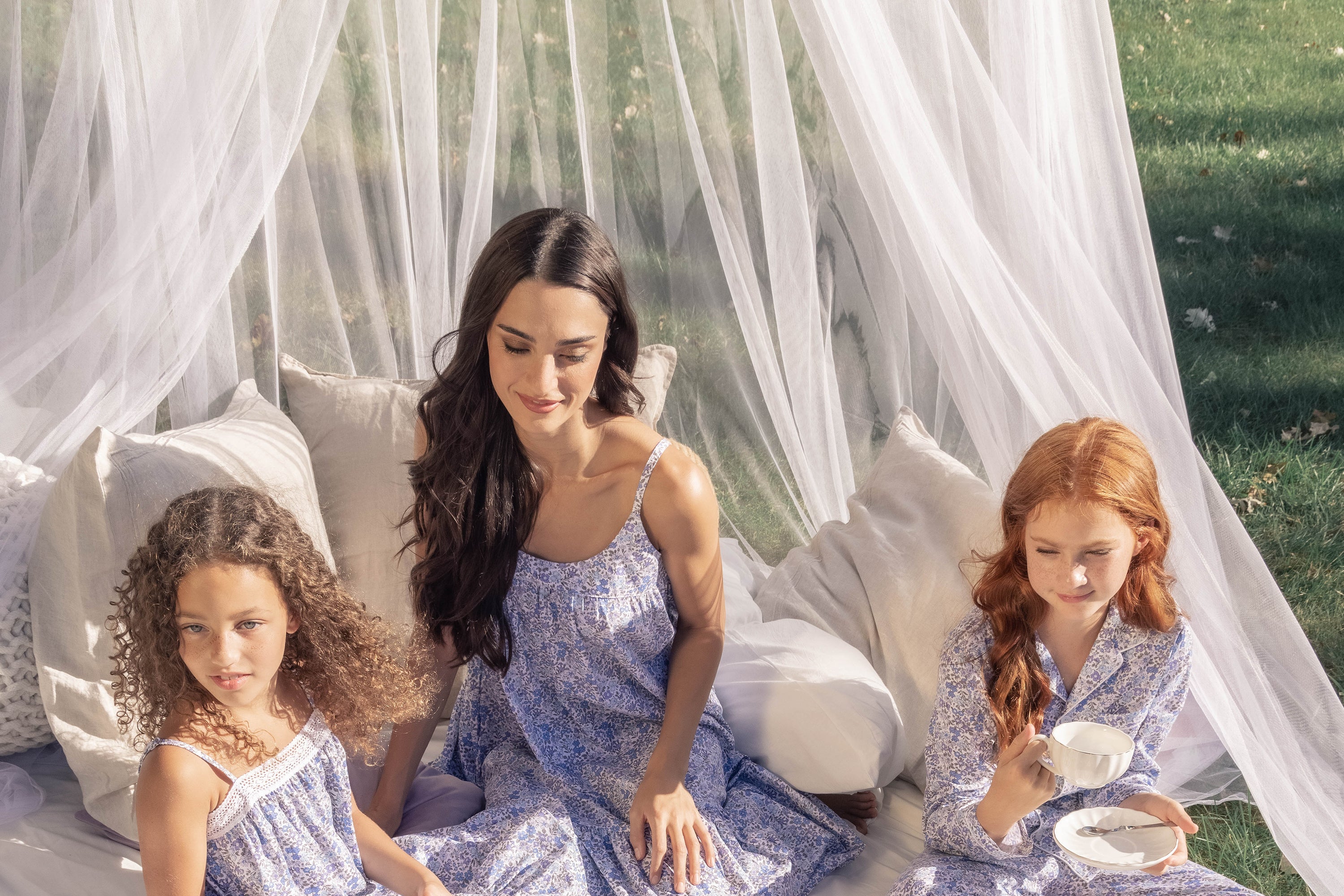 A woman and two young girls in matching light purple floral dresses sit on a bed outdoors under a sheer white canopy; one girl pours tea while the others look on, surrounded by pillows and greenery.