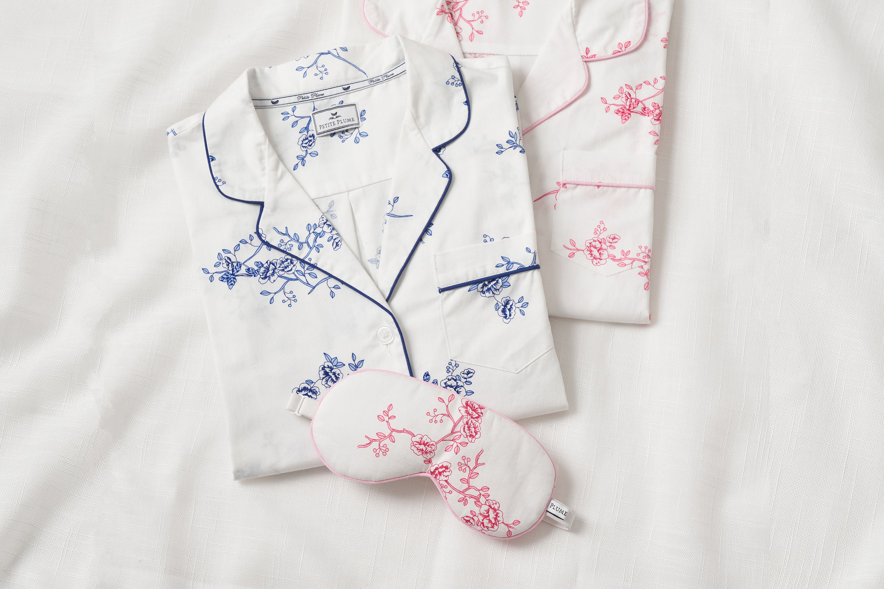Two sets of folded pajamas with floral patterns, one blue and one pink, are laid on a white fabric background. A matching pink-trimmed sleep mask is placed in front of the pink pajama set.