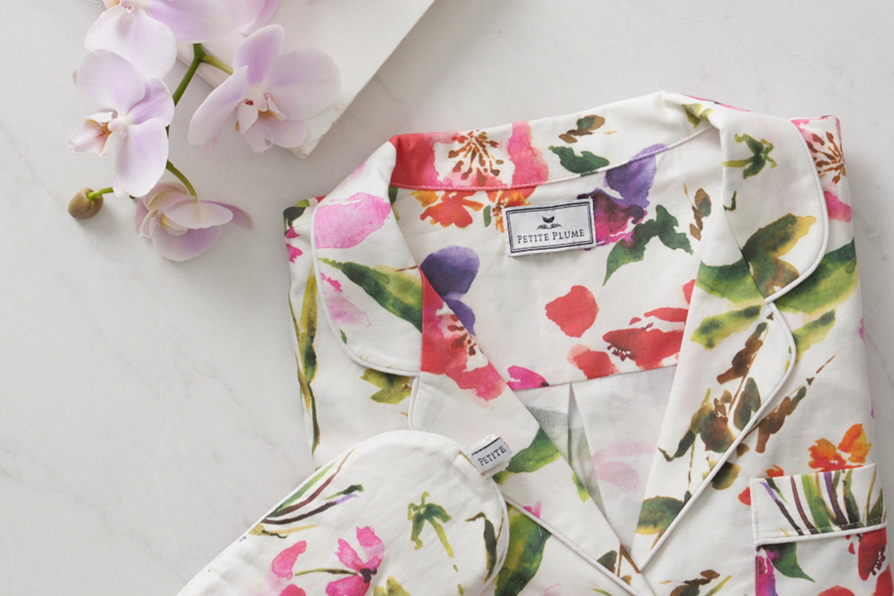 Floral-print pajamas with green, pink, and red patterns are neatly folded on a white marble surface next to light pink orchids and a white envelope.