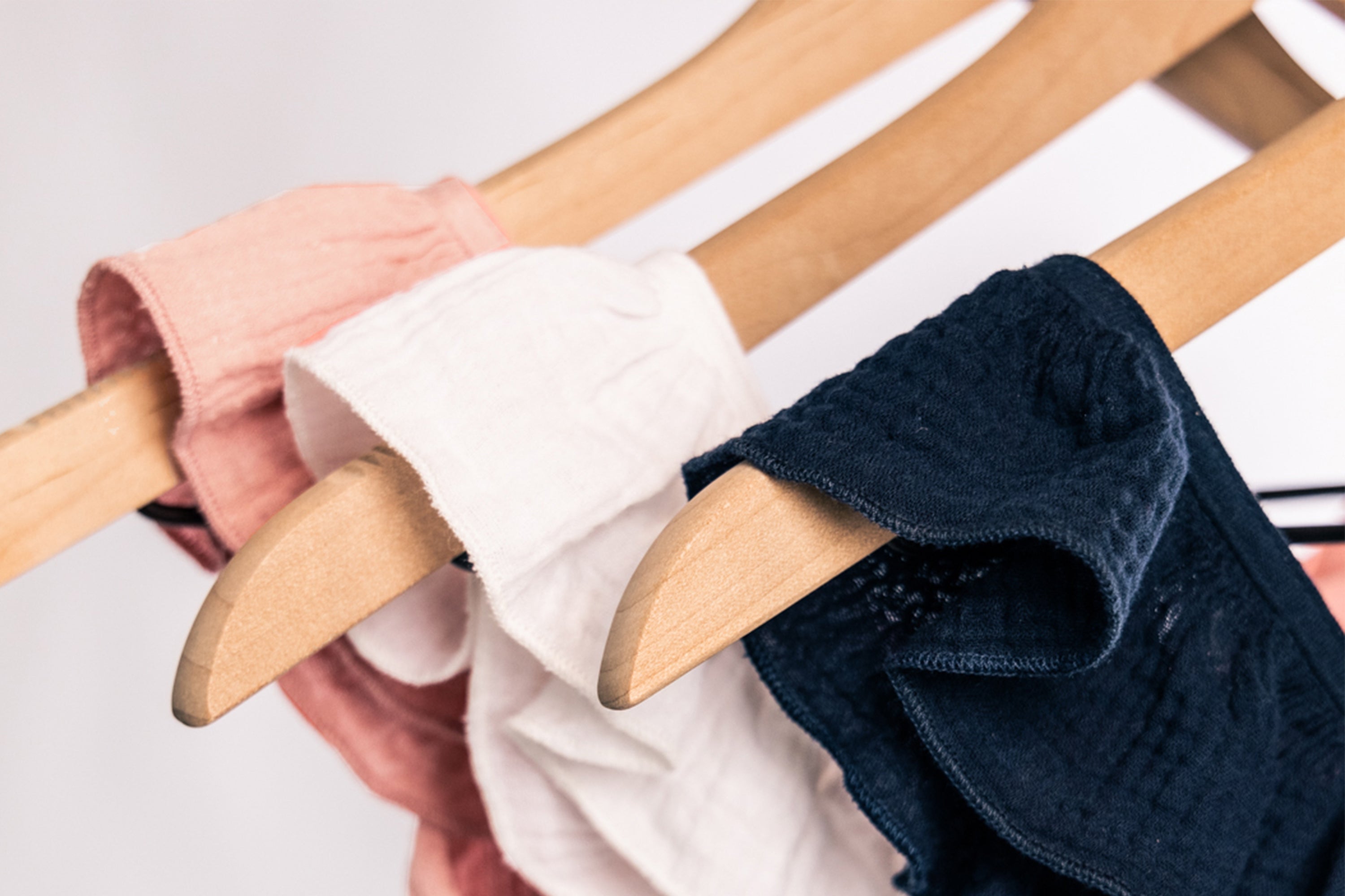 Three wooden hangers hold clothing items in soft pink, white, and dark navy blue fabrics, displayed against a neutral background. The clothes appear to be lightweight and casual.