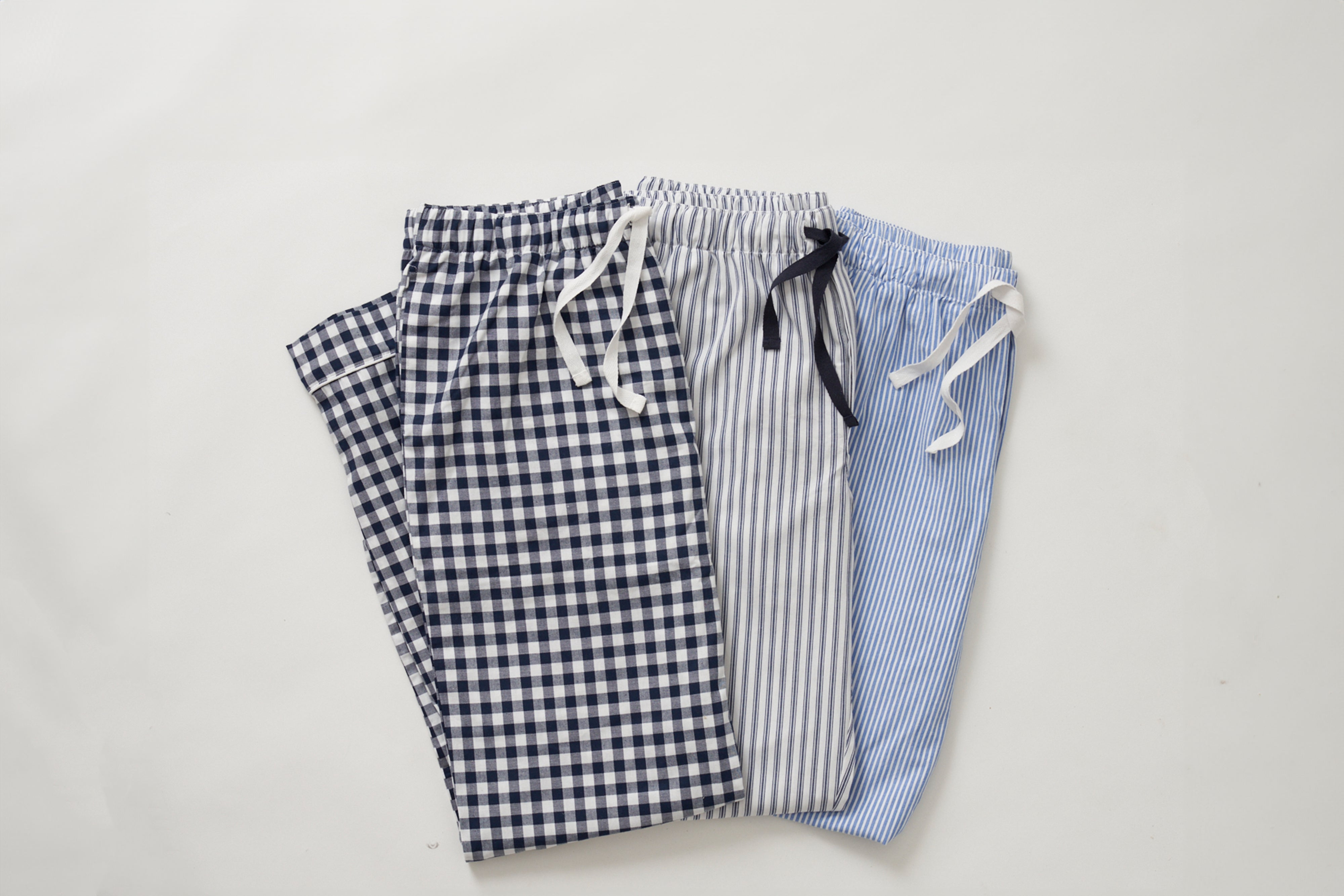 Three pairs of pajama pants on a white background. The pants have different patterns: dark blue and white checkered, gray and white striped, and light blue and white striped. Each has a drawstring.