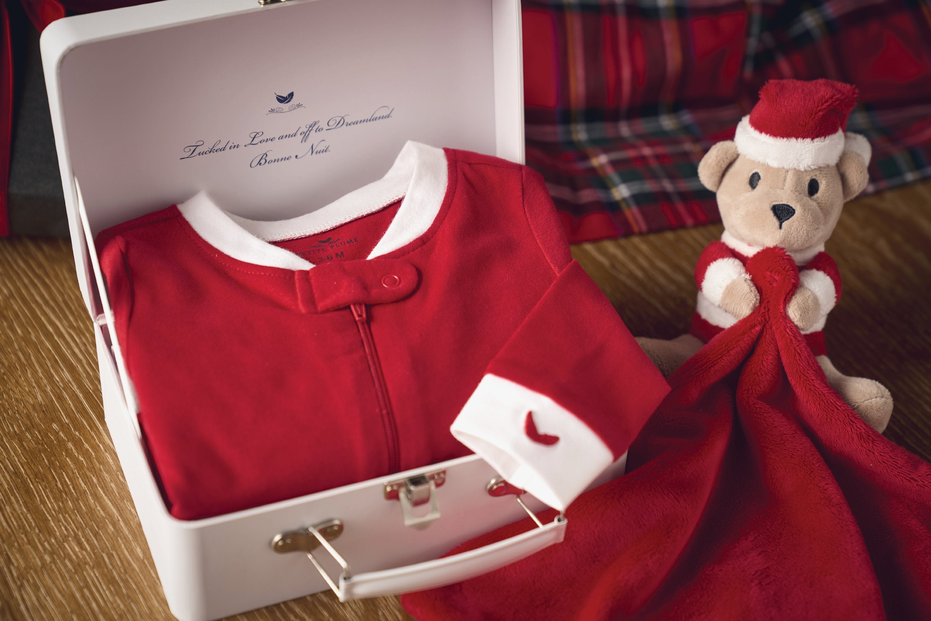 A red baby outfit with white trim is neatly placed in an open gift box. Next to it, a plush teddy bear wearing a Santa hat holds a red blanket. The background features a plaid fabric.
