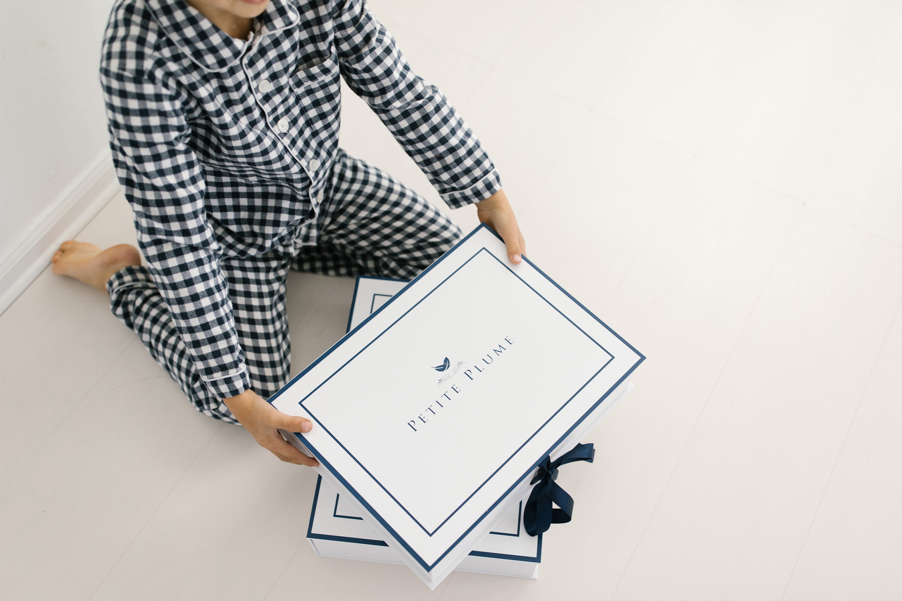 A child in checkered pajamas sits on the floor, holding a large white box labeled Petite Plume with a blue border and logo, with another similar box beneath it.