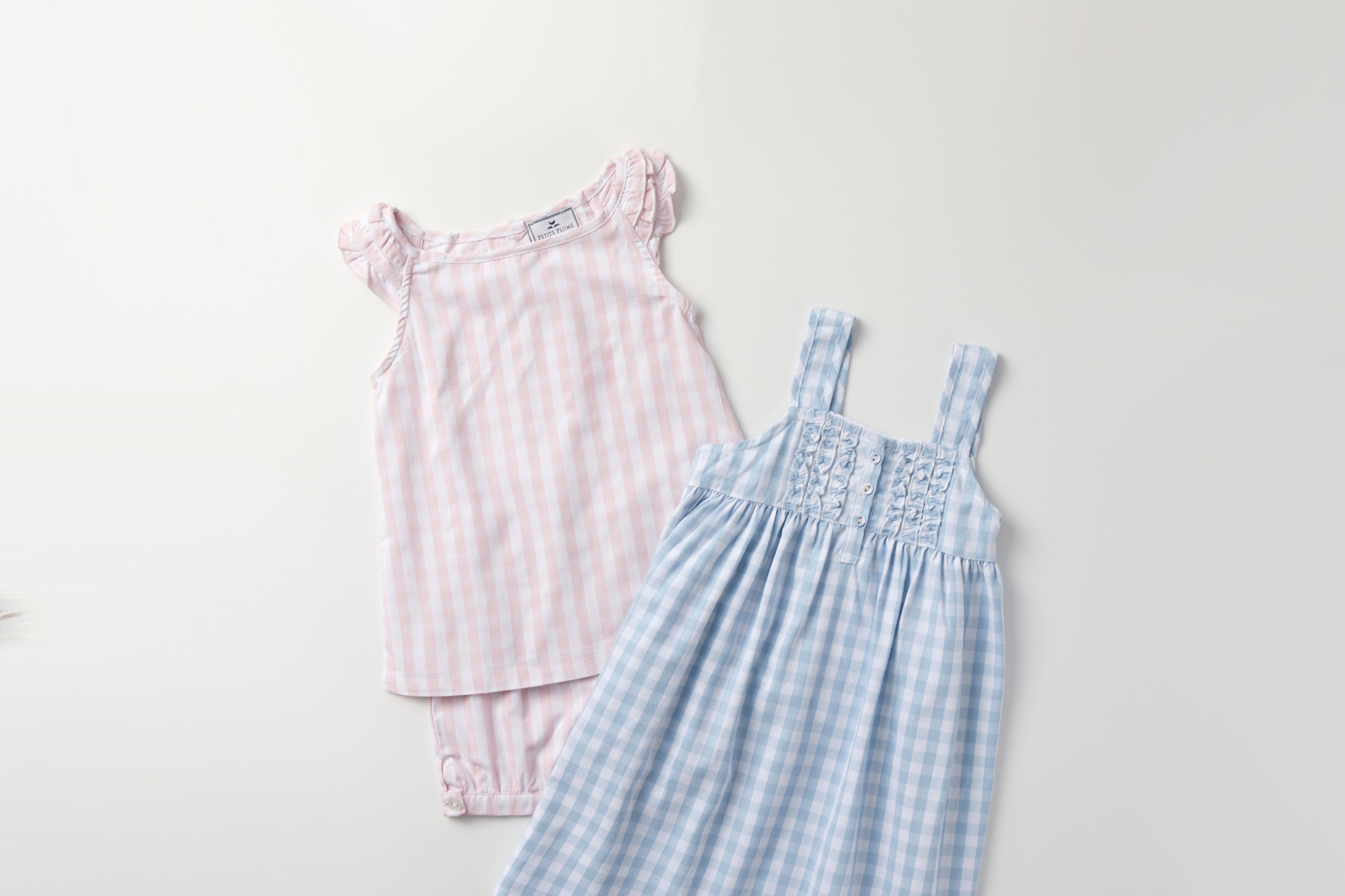 Two childrens outfits on a white background: a pink gingham sleeveless top with matching shorts, and a light blue gingham sundress with straps and lace detail on the bodice.