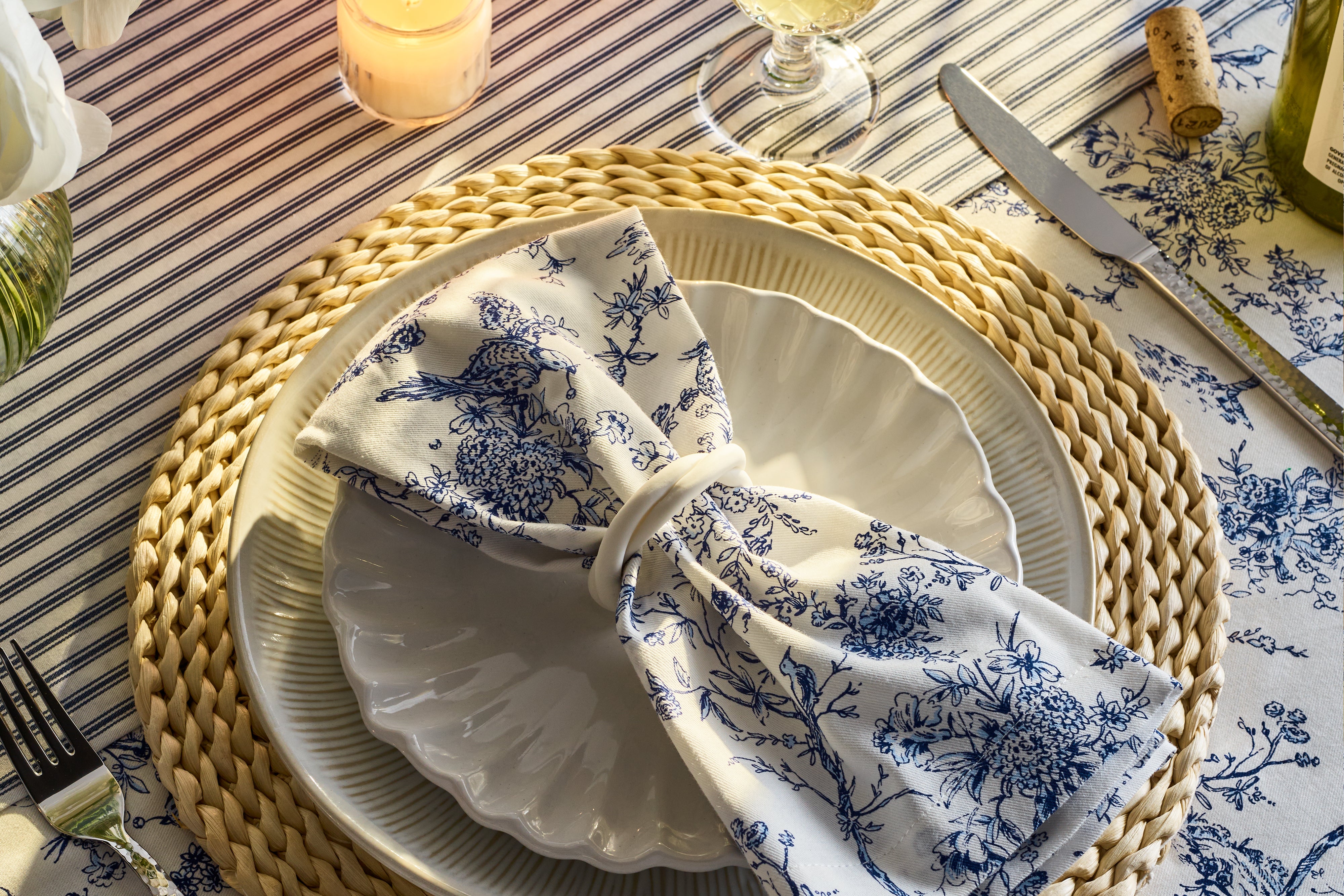 A place setting with a woven placemat, white plates, a blue floral napkin in a ring, silverware, a wine glass, a lit candle, and a cork, all on a blue and white striped tablecloth.