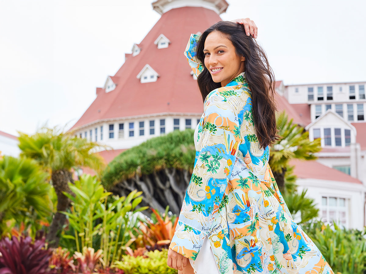 A woman in a colorful, tropical-patterned dress smiles while turning back, standing in front of a historic, red-roofed building surrounded by greenery and flowers.