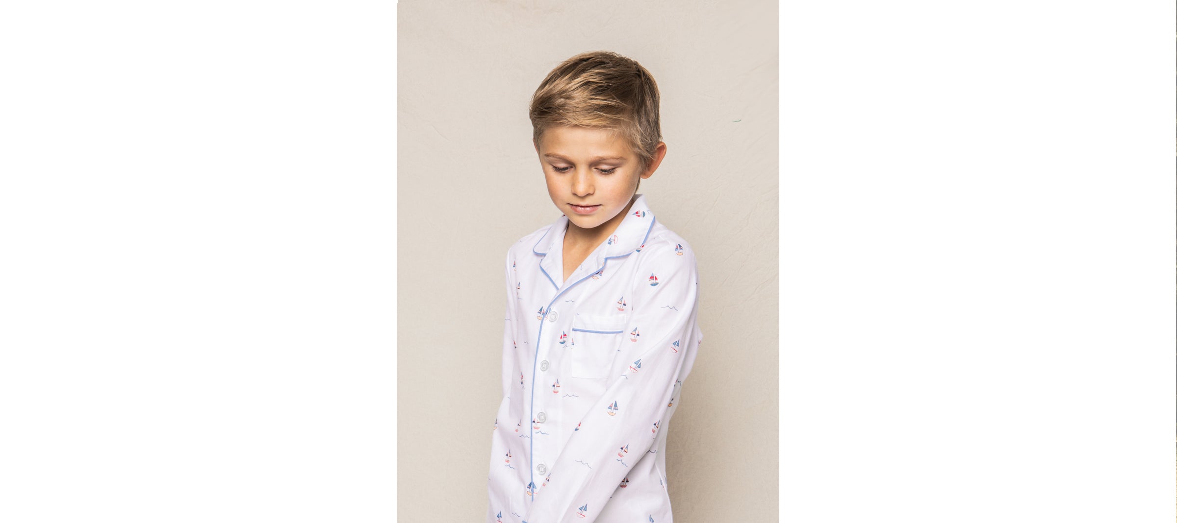 A child with short blond hair, wearing white pajamas with small patterns, stands against a plain background. The pajamas have a pocket and blue trim. The child looks downwards with a slight smile.
