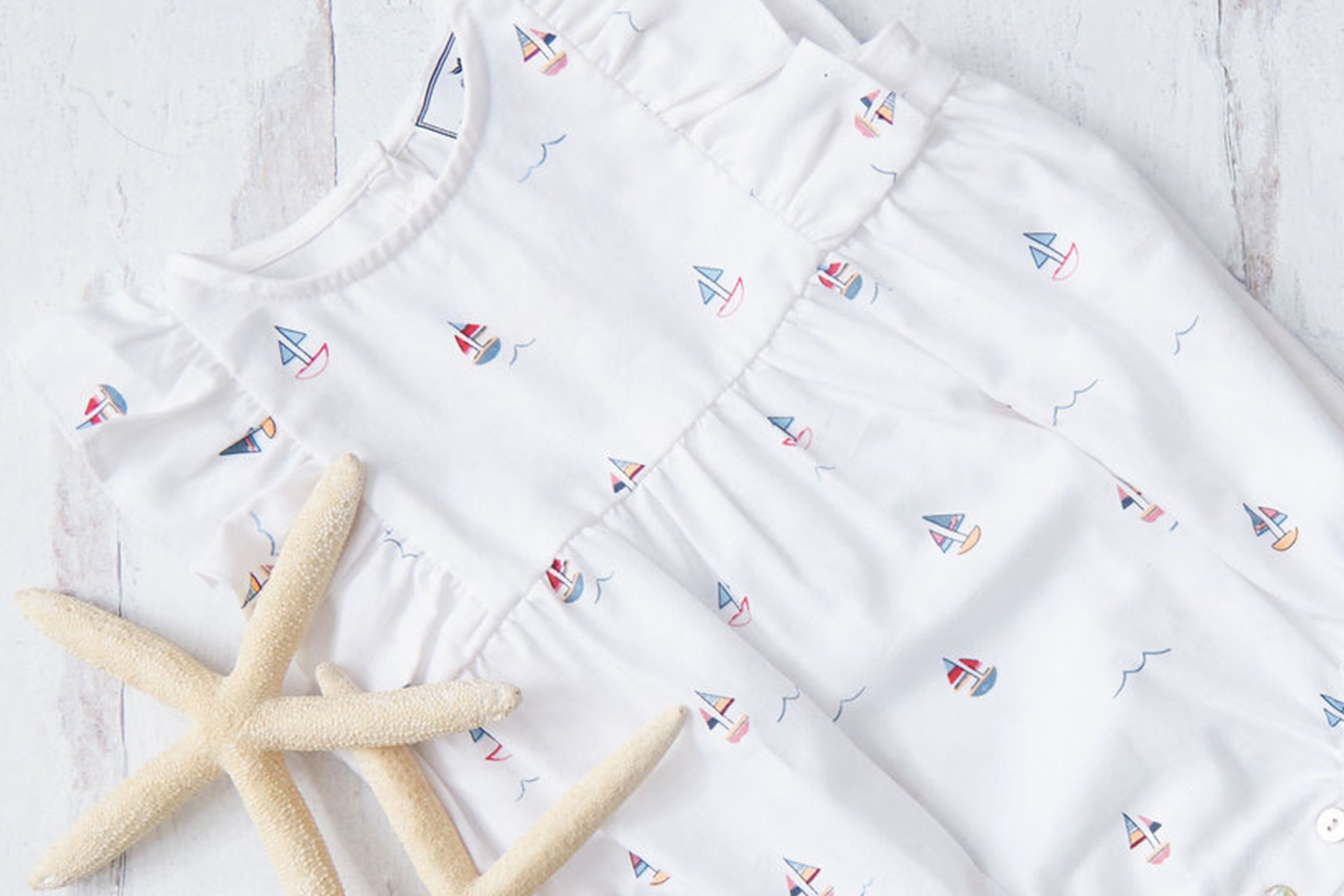 A white baby romper with colorful sailboat prints is laid flat on a light wooden surface beside two beige starfish.