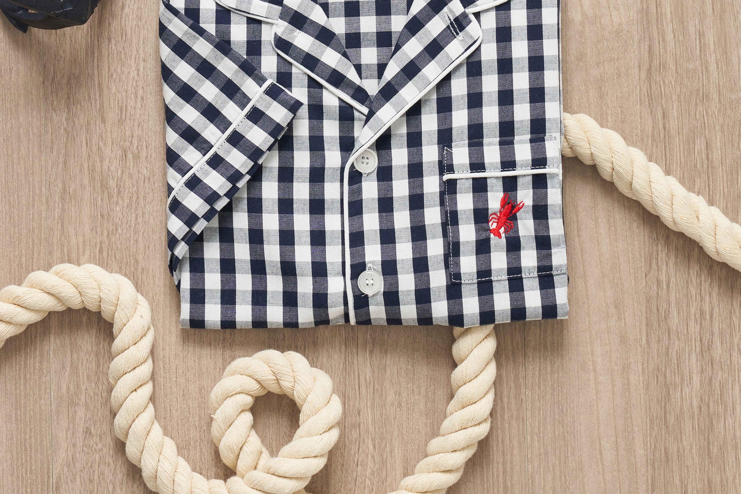 A folded navy and white checkered pajama shirt with white piping and a red lobster embroidered on the pocket, placed on a wooden surface with thick, coiled rope.