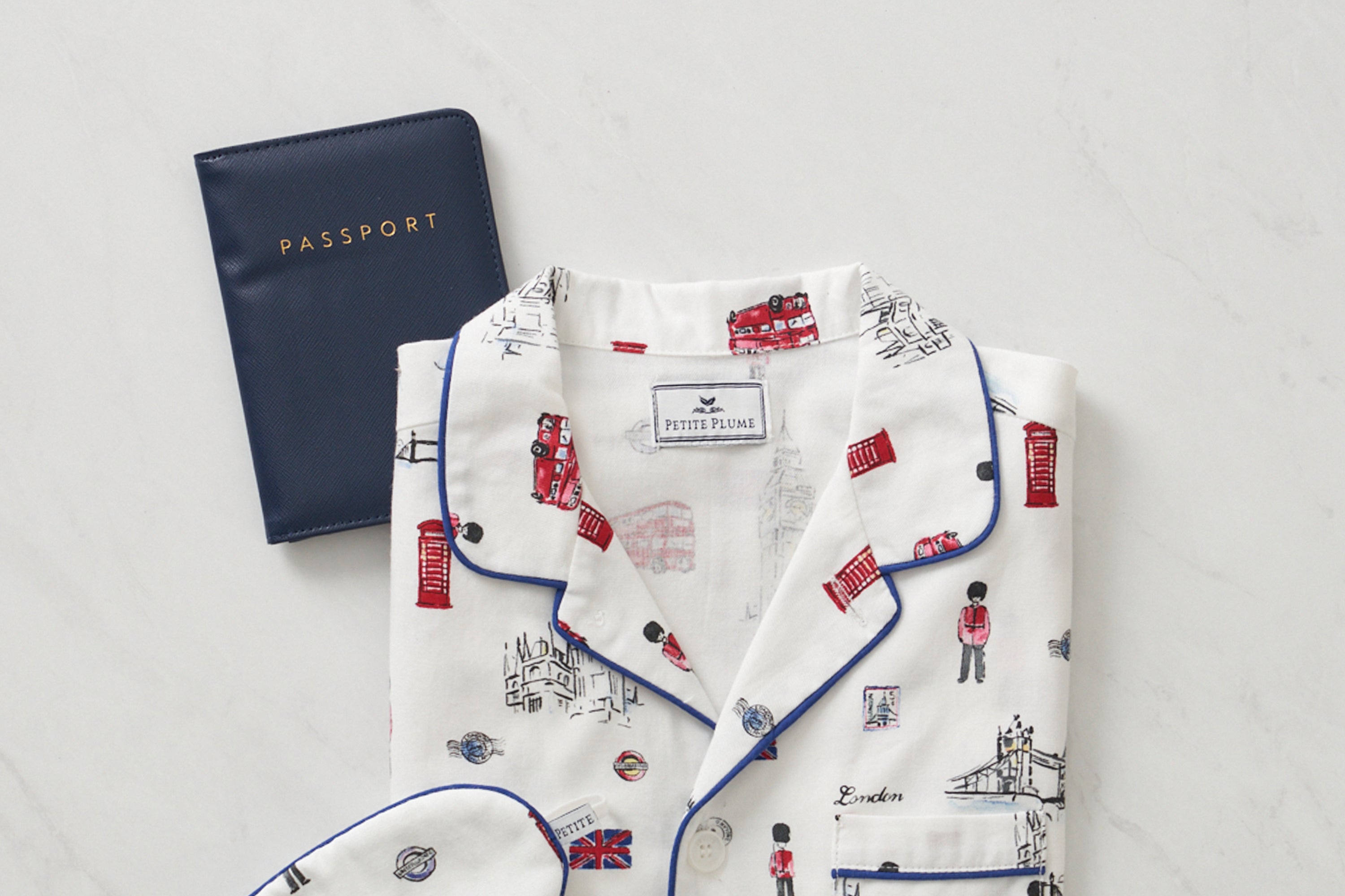 A folded white pajama shirt with London-themed prints sits next to a closed navy blue passport on a light marble surface.