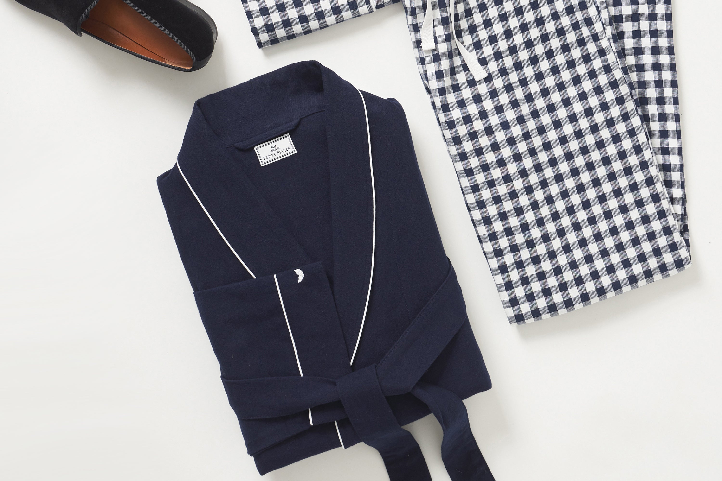 A folded navy blue bathrobe with white piping, next to blue and white checkered pajama pants, and a partial view of a black slipper on a light background.