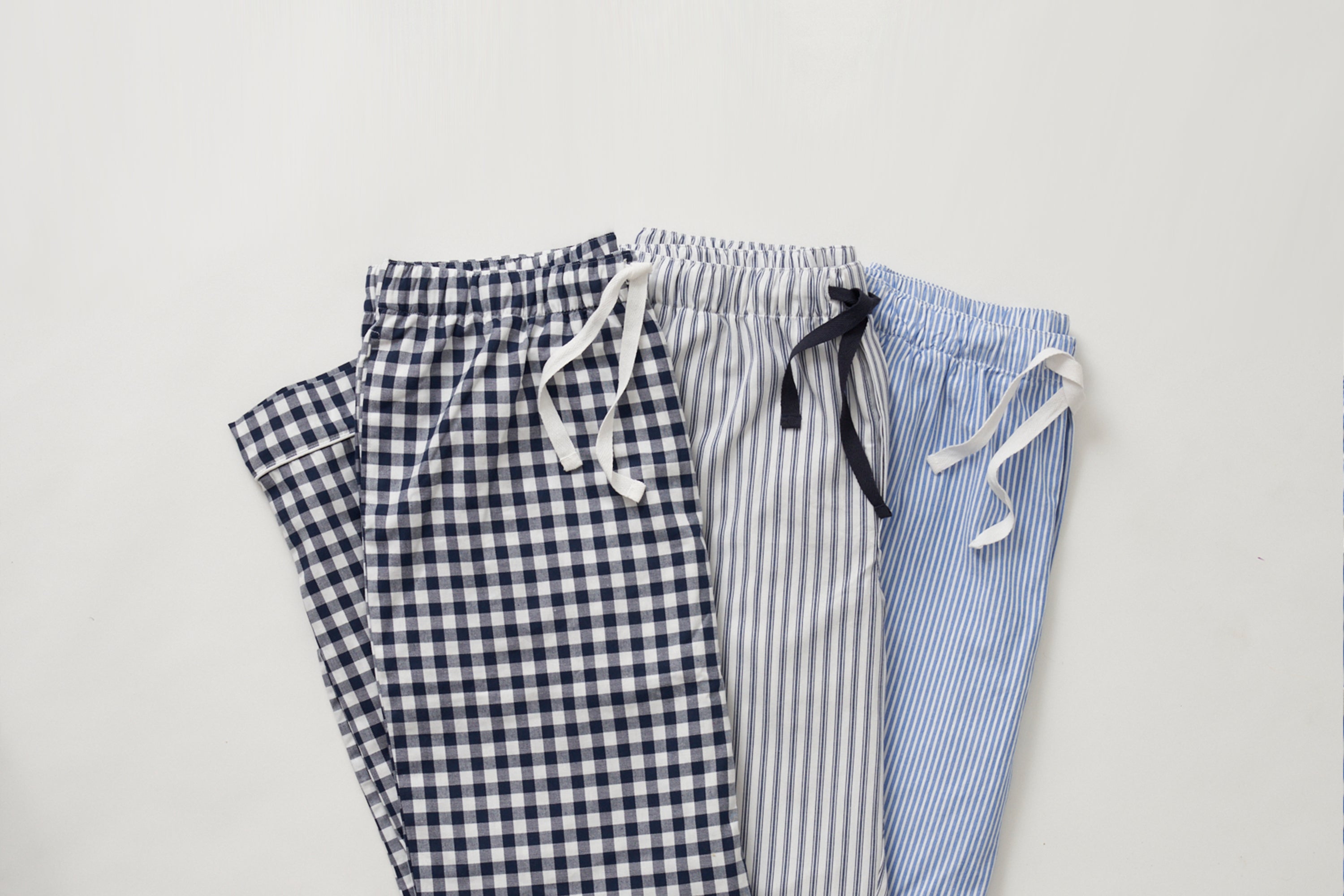 Three pairs of pajama pants with drawstrings are laid out side by side: one with a blue checkered pattern, one with gray vertical stripes, and one with thin blue and white pinstripes.
