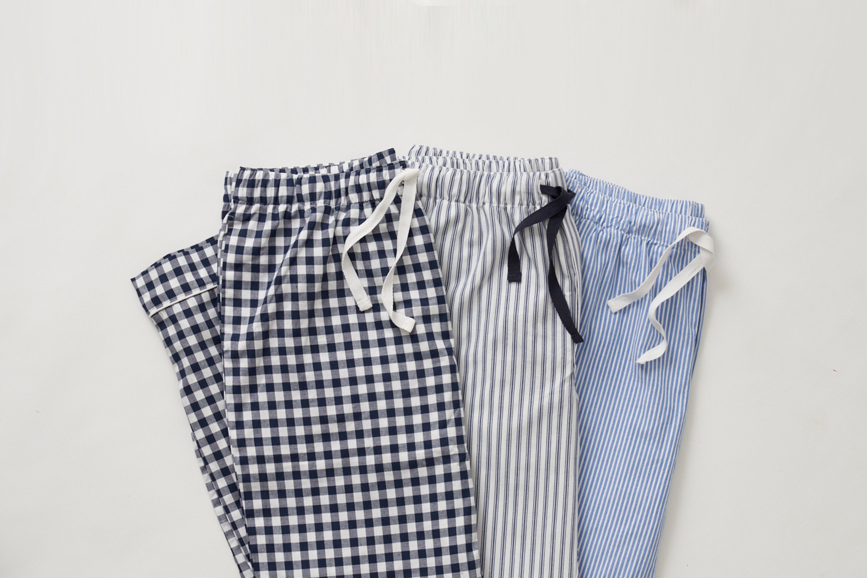 Three pairs of drawstring pajama pants are laid side by side. The pants feature different patterns: navy gingham, gray pinstripe, and light blue pinstripe, all on a white background.