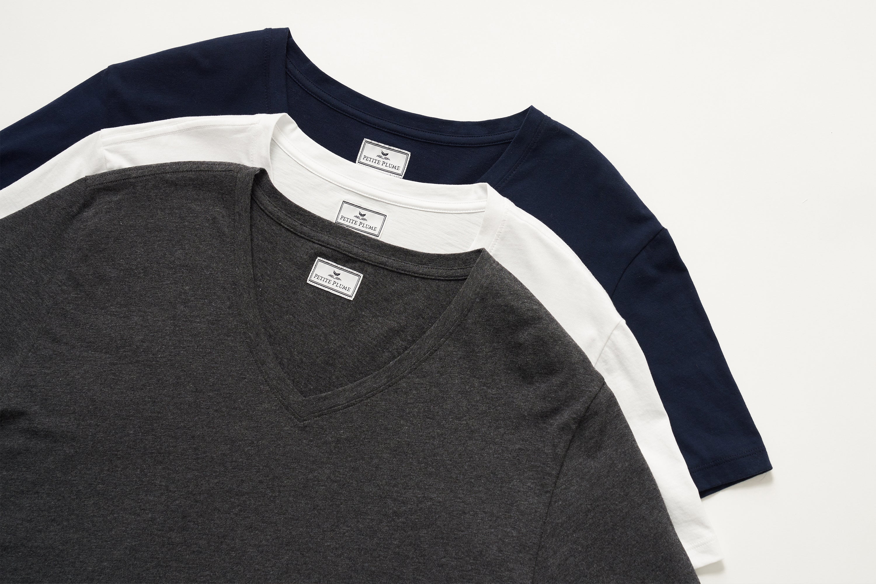 Three neatly folded V-neck T-shirts in dark gray, white, and navy blue are stacked on a white surface, each with visible labels at the collar.