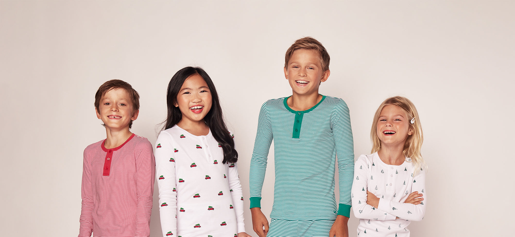 Four children stand smiling against a plain background, wearing festive pajamas in red, green, and white with striped and holiday-themed patterns.