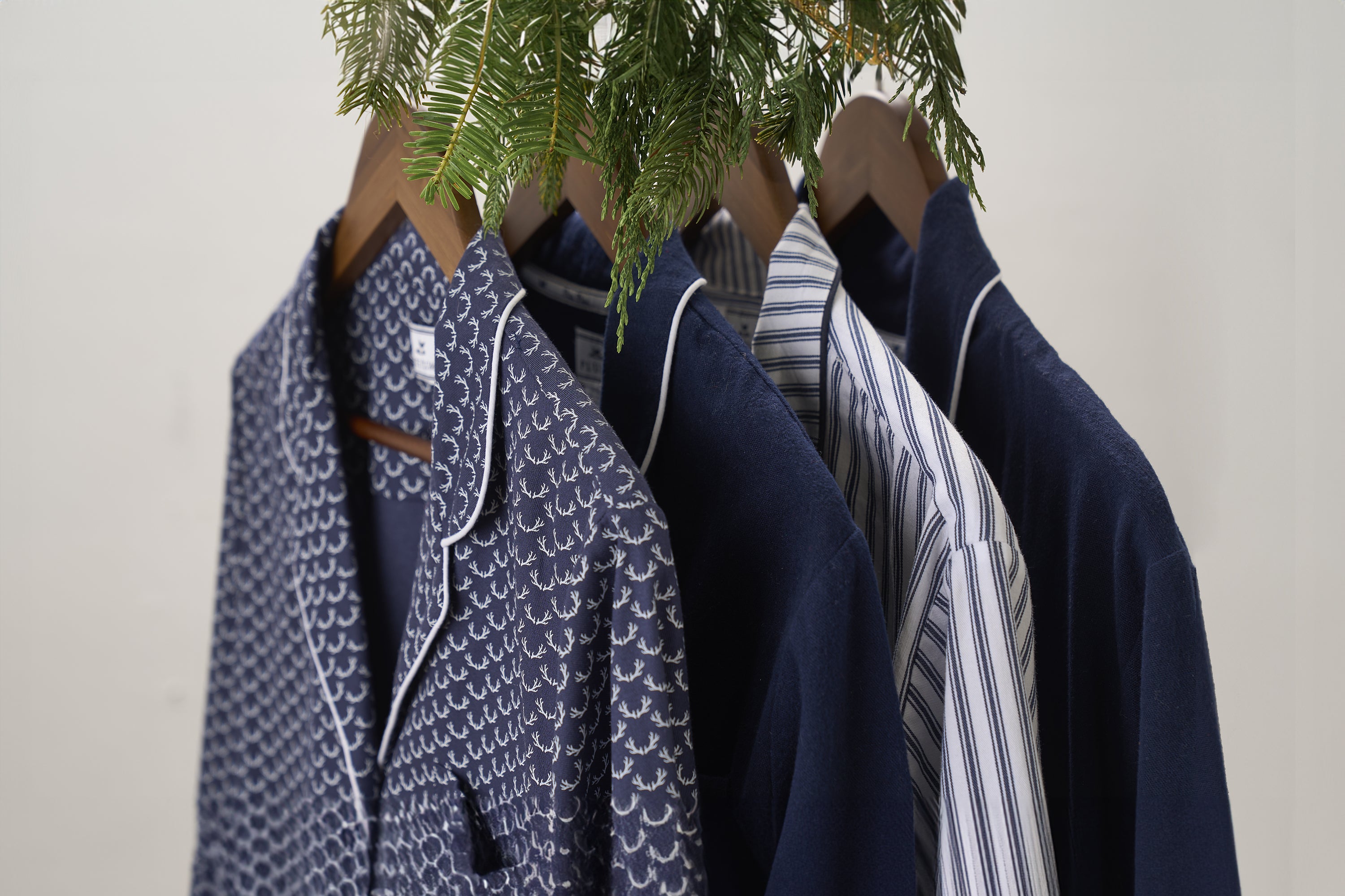 Four shirts on wooden hangers, including a patterned navy pajama top and three button-up shirts in solid navy and striped designs, hang beneath green foliage.