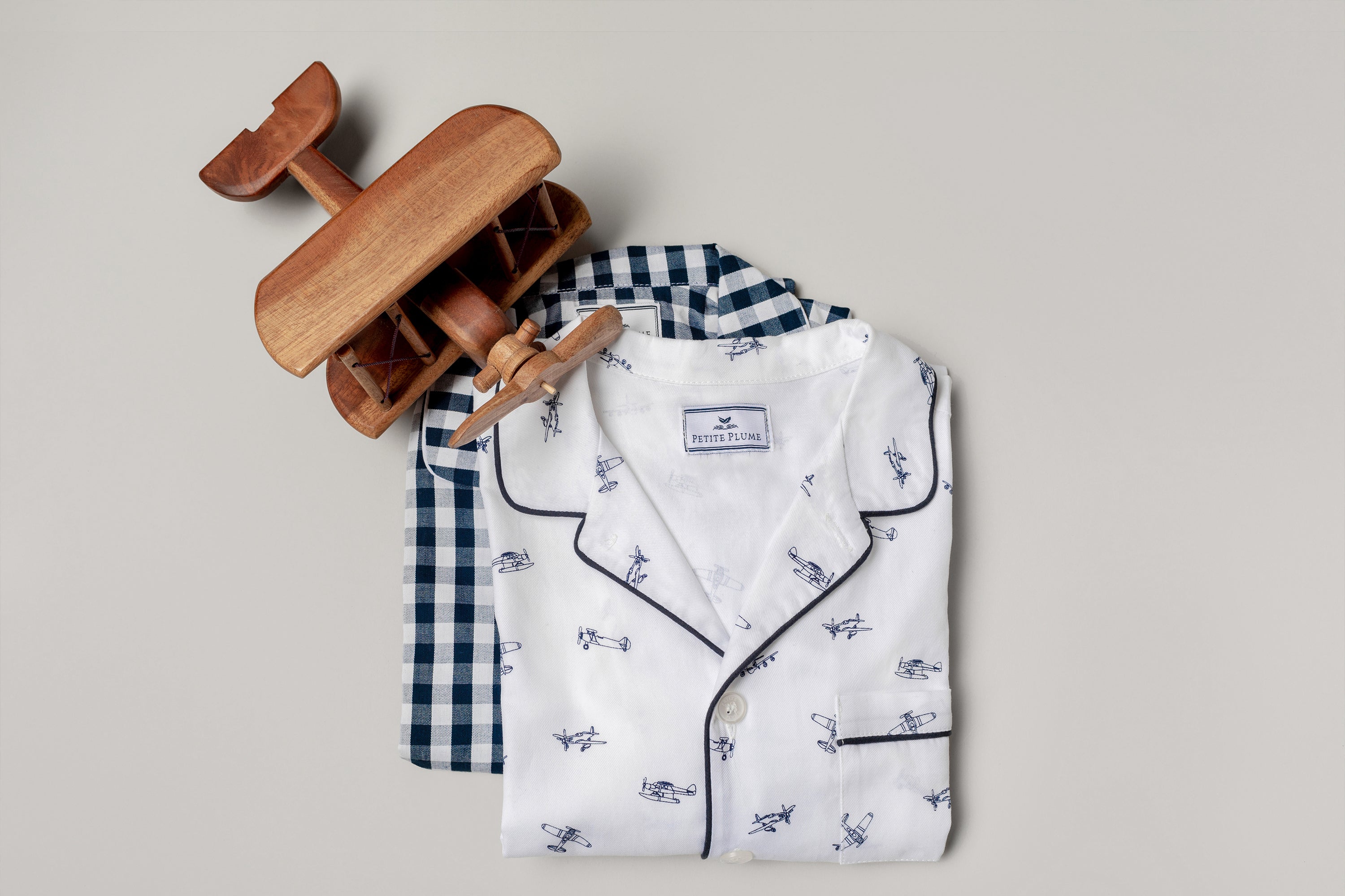 Two sets of folded children’s pajamas, one blue gingham and one white with airplane prints, are stacked next to a small wooden toy airplane on a light gray background.