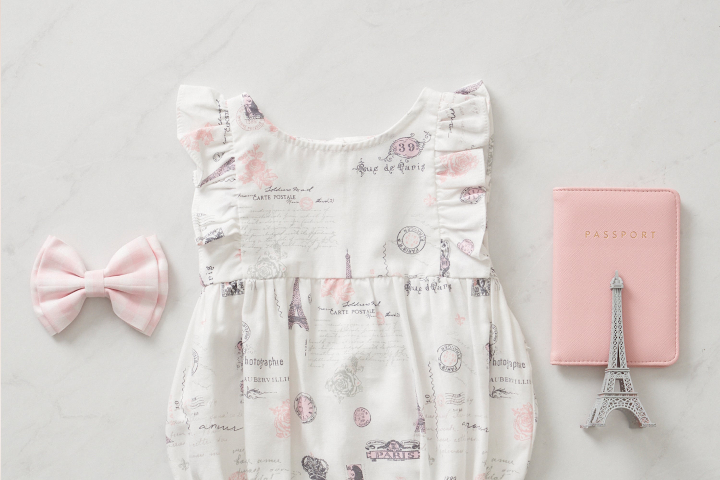 Flat lay of a baby dress with Paris-themed prints, a pink hair bow, a miniature Eiffel Tower, and a pink passport cover arranged on a light surface.