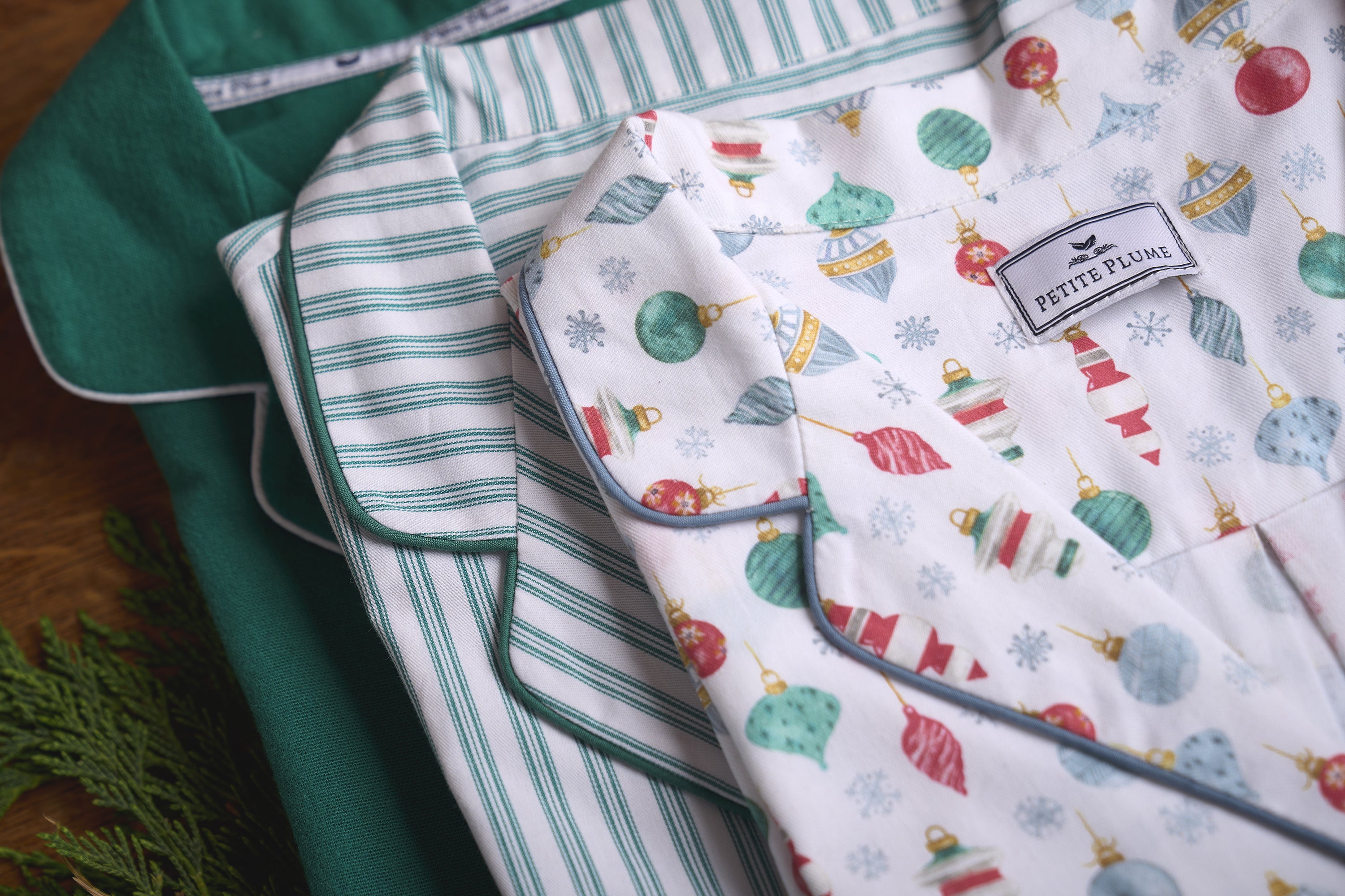 Three folded pajamas are stacked: the top has a festive holiday print with ornaments, the middle has green and white stripes, and the bottom is solid green. A pine branch is partially visible in the corner.