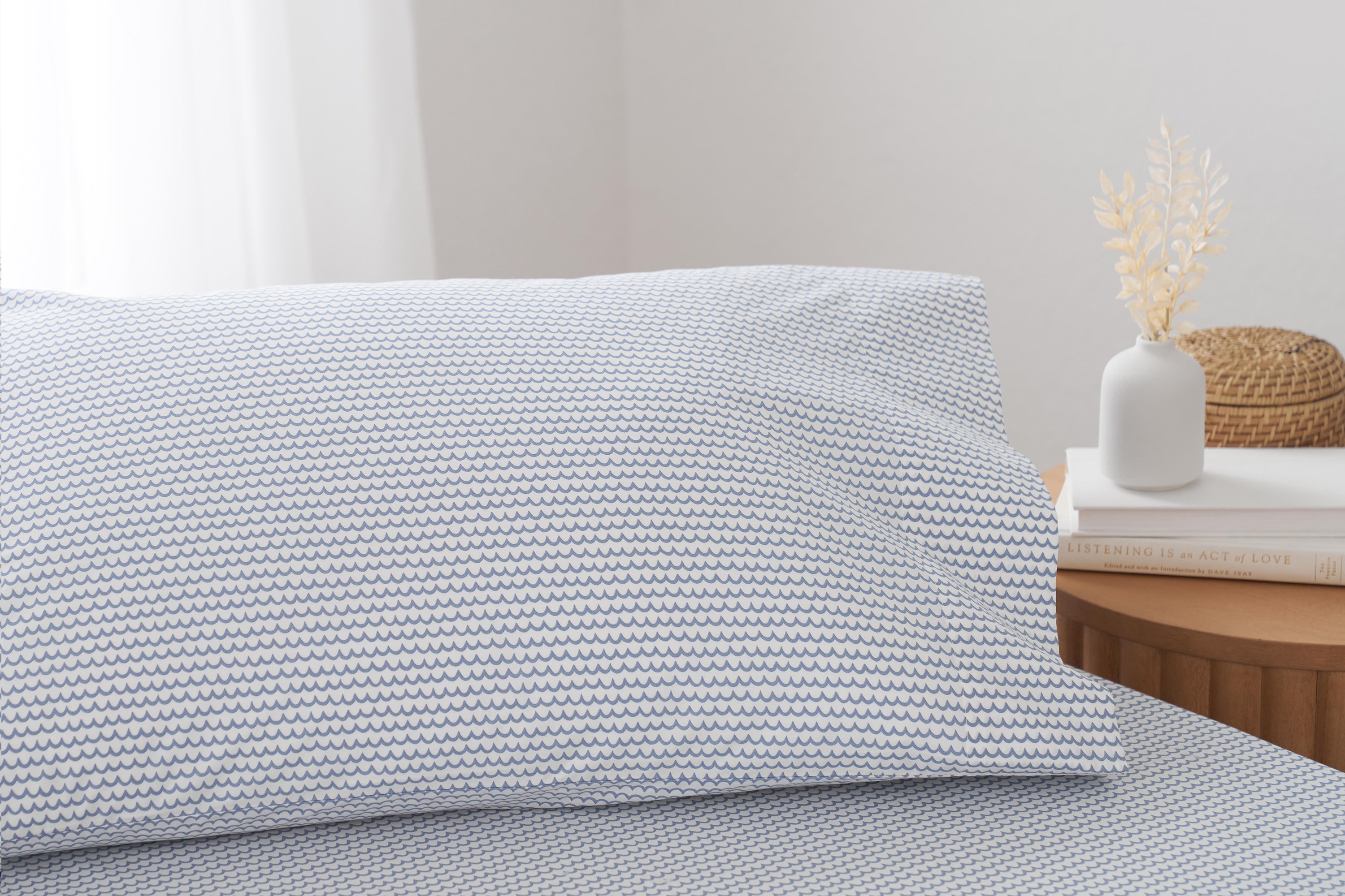 A close-up of a bed with a pillow and sheet featuring a small blue and white zigzag pattern. Next to the bed is a wooden nightstand with books, a white vase, and dried flowers.